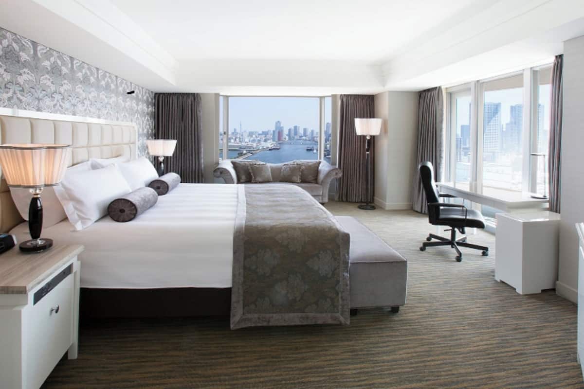 A hotel room with a large bed, a desk, a chair, a lamp, and a view of the cityscape.
