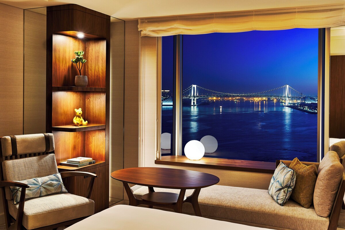 Una habitación moderna con un ventanal grande que da a un puente de noche, un sofá, una silla y una mesita.