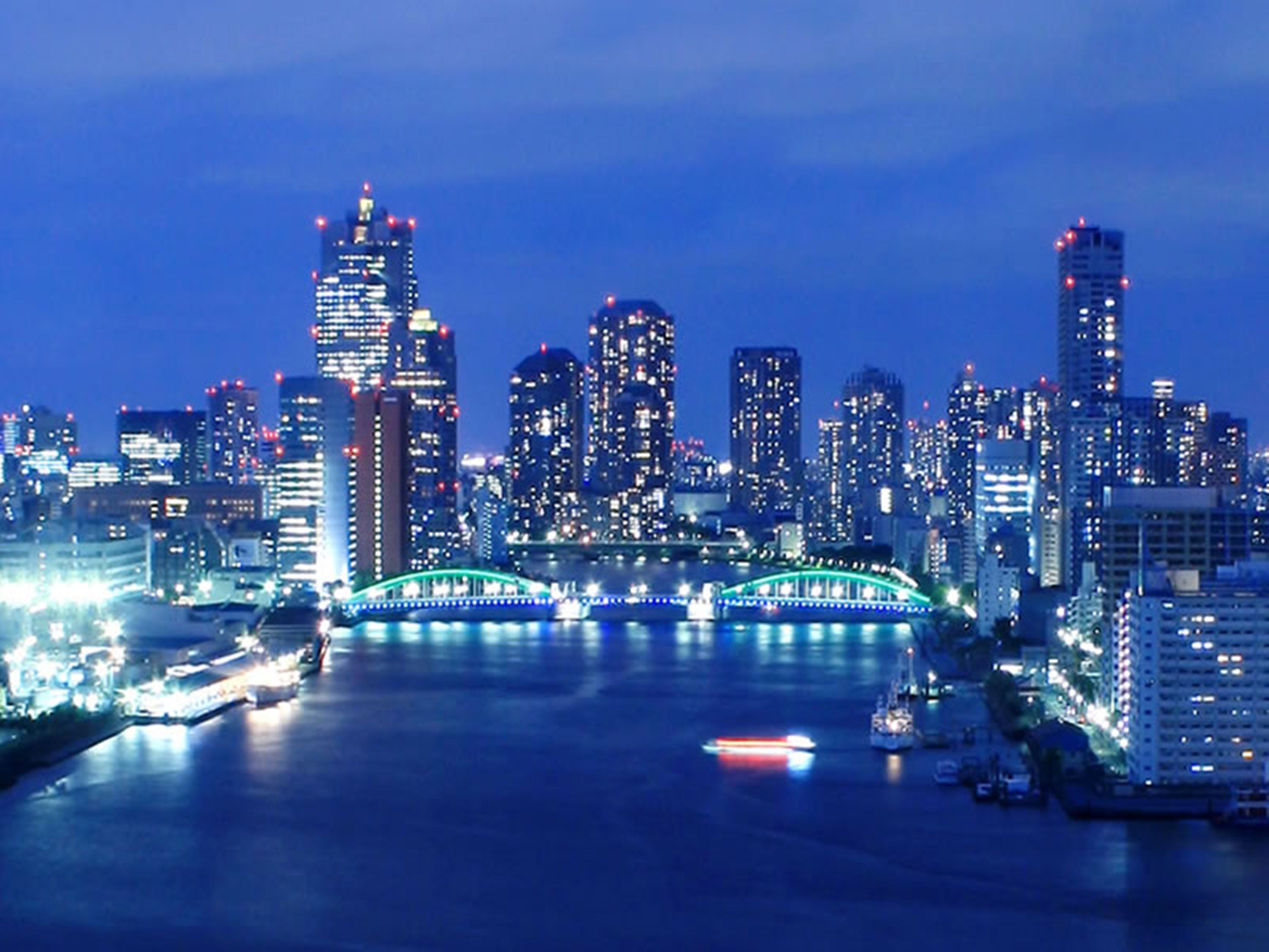 A nighttime cityscape with illuminated skyscrapers and a bridge over a river.