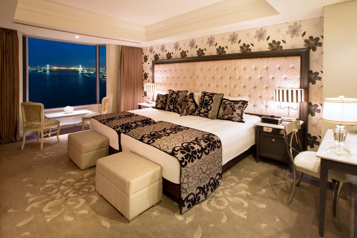 A hotel room with a large bed, a desk, a chair, and a view of a cityscape at night.