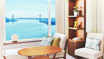 A room with a large window overlooking a bridge and water, a wooden table, two armchairs, and a bookshelf with decorative items.