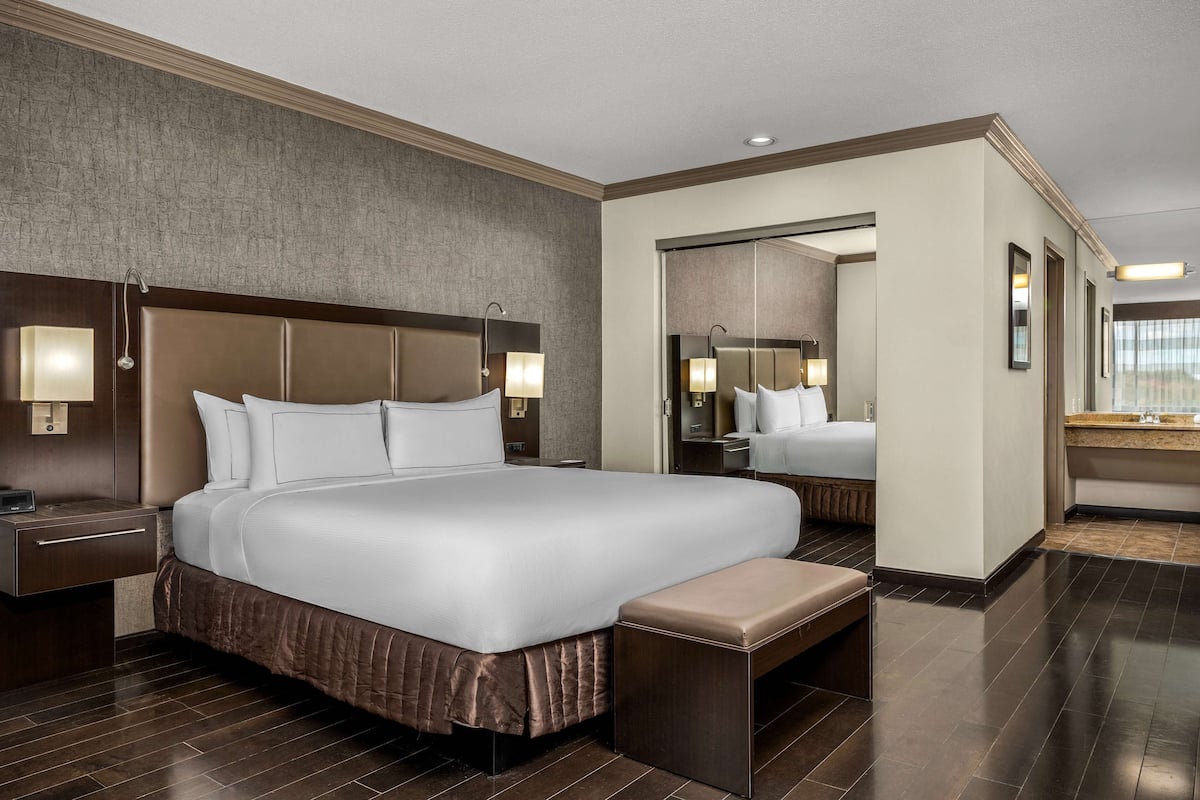 A modern hotel room with a large bed, bedside tables, a bench, a mirror, and a bathroom with a sink and mirror.