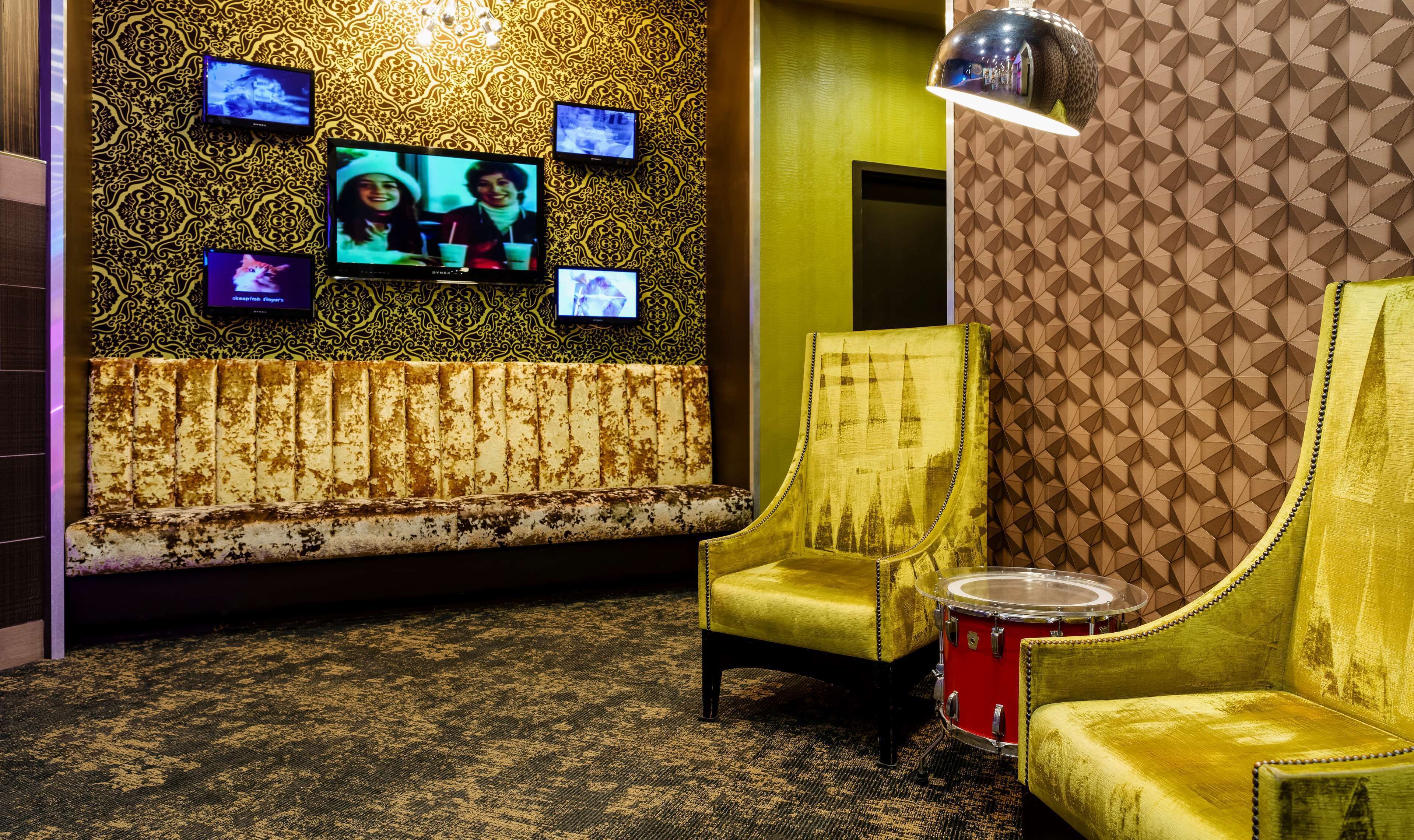 A room with a patterned wall, a sofa, two armchairs, a drum set, and multiple screens.