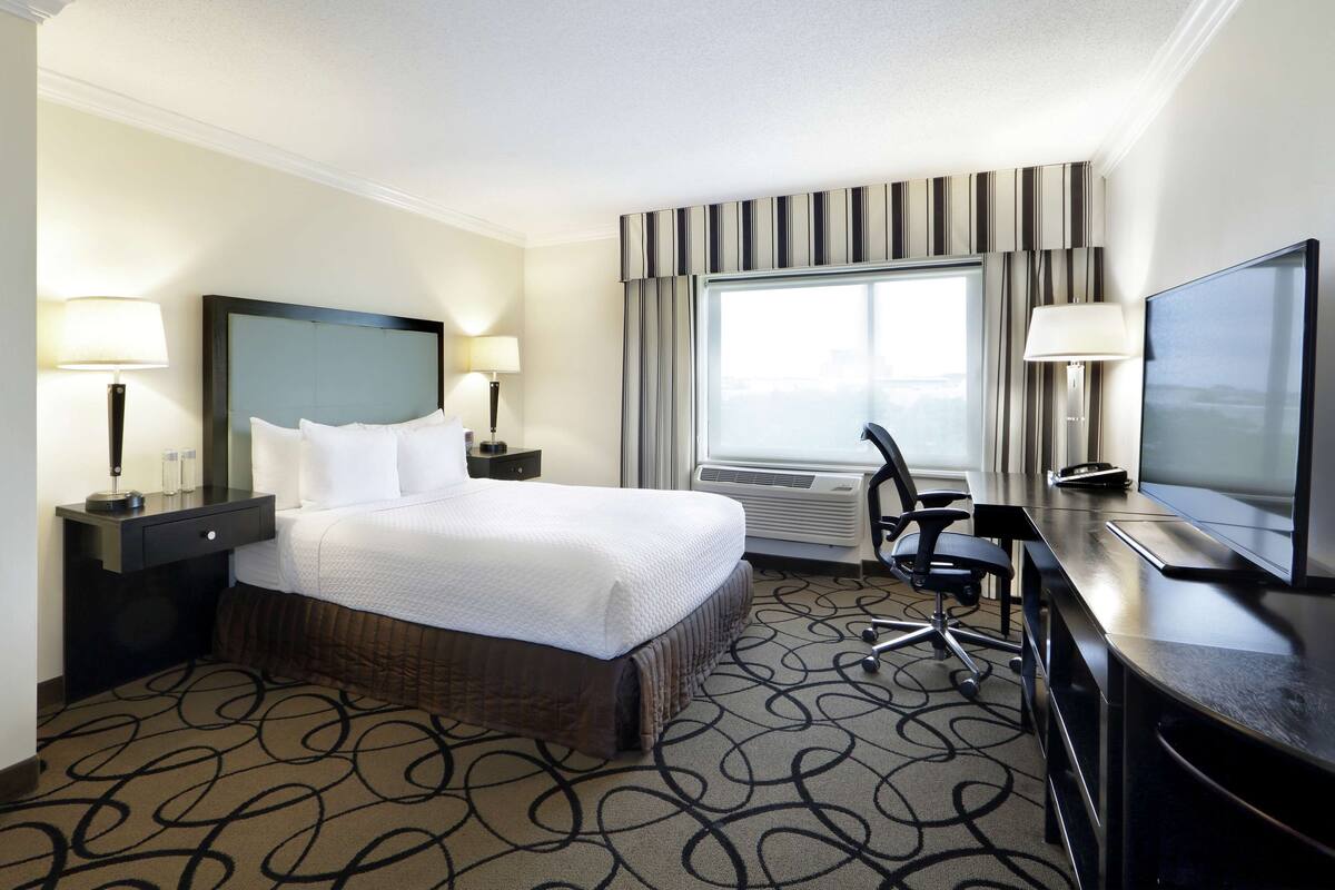 A hotel room with a large bed, bedside tables with lamps, a desk with a chair, a television, and a large window with curtains.