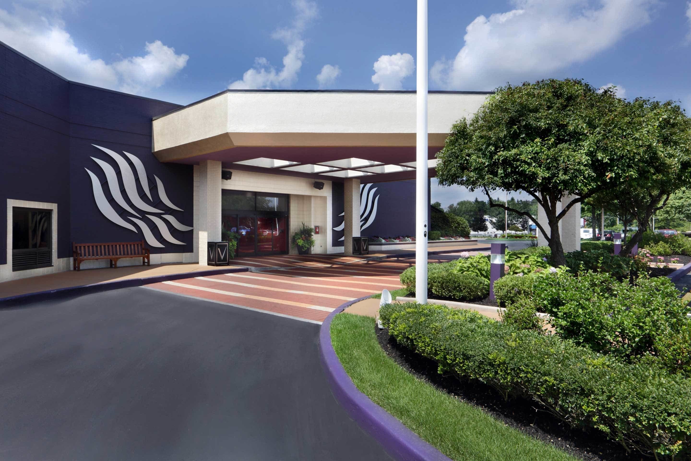 A modern building with a large entrance, a covered waiting area, and a landscaped garden with a tree and bushes.