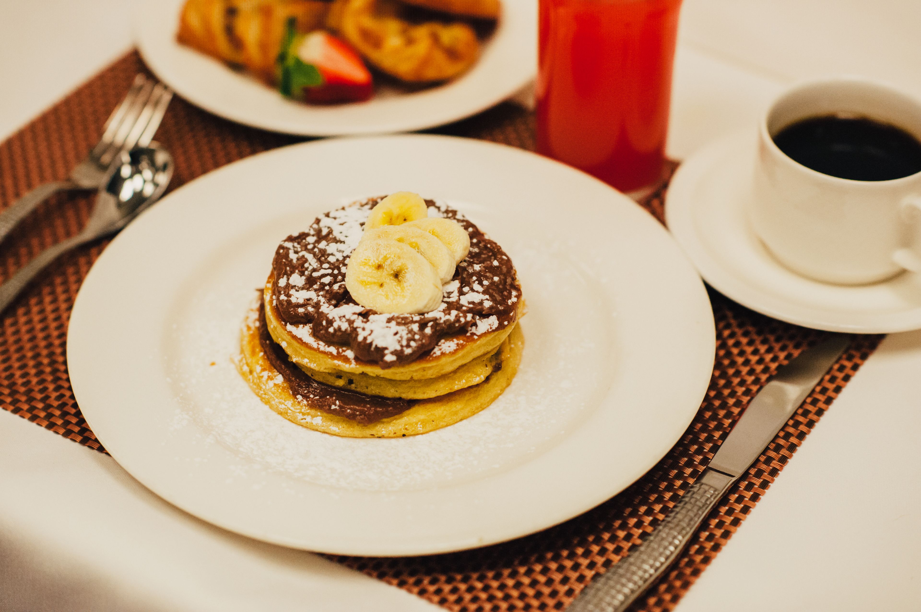 A breakfast setting with pancakes topped with bananas and powdered sugar, a cup of coffee, and a croissant on a plate.