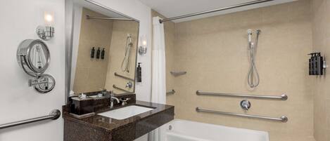 A modern bathroom with a sink, mirror, and bathtub. It includes grab bars and a shower with a handheld showerhead.