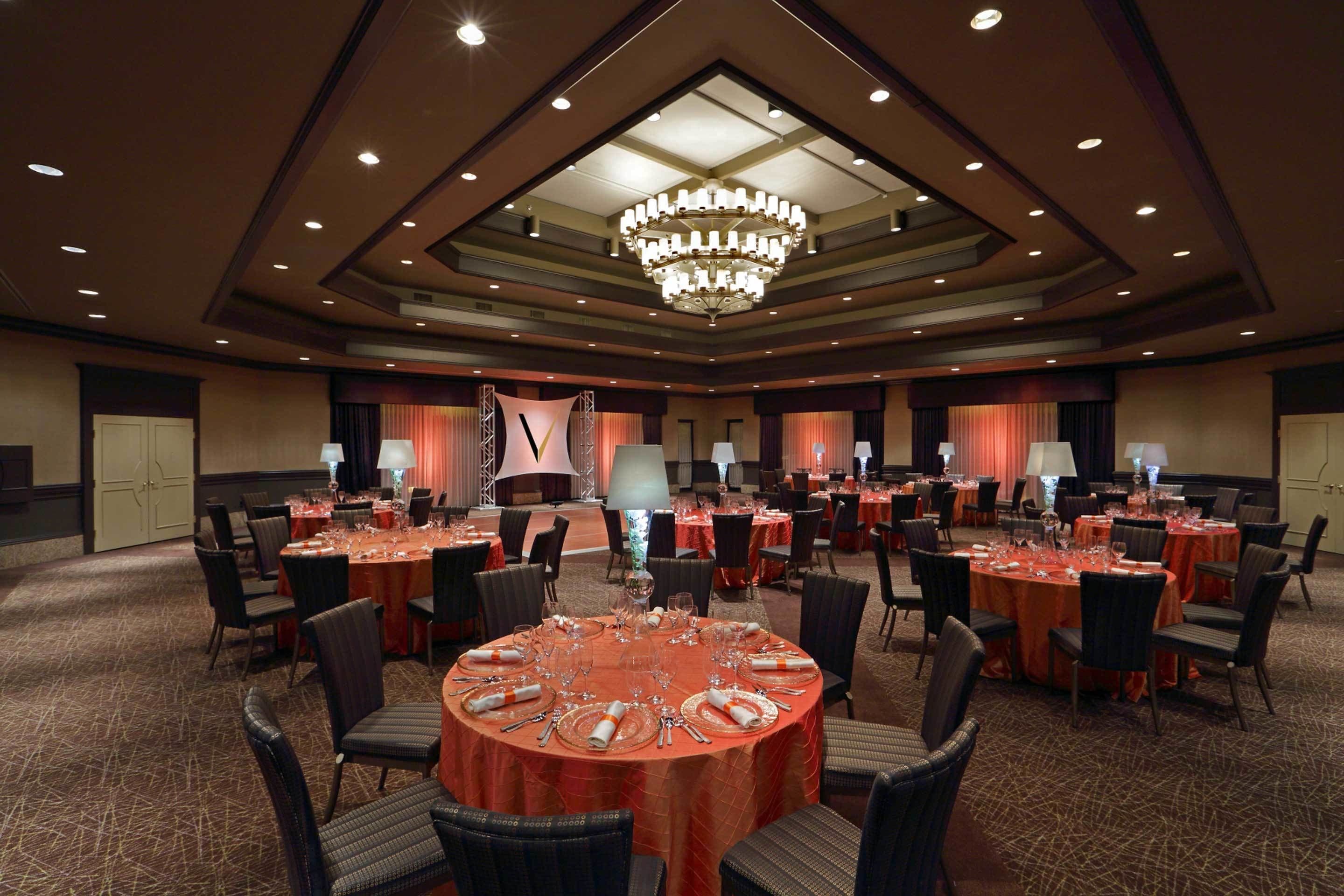 A large conference room with multiple round tables, each set with plates, cutlery, and glasses. The room features a high ceiling with recessed lighting and a central chandelier.