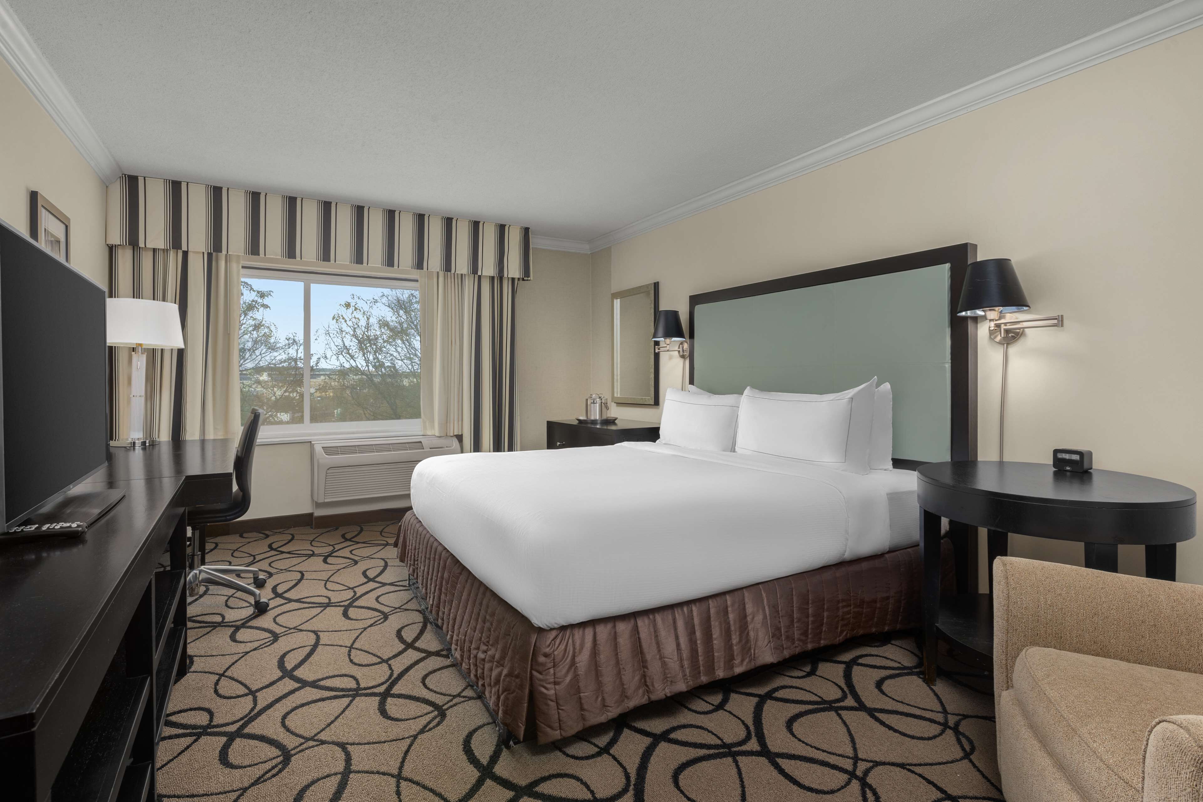 A hotel room with a large bed, a desk with a chair, a TV, a lamp, and a window with curtains.