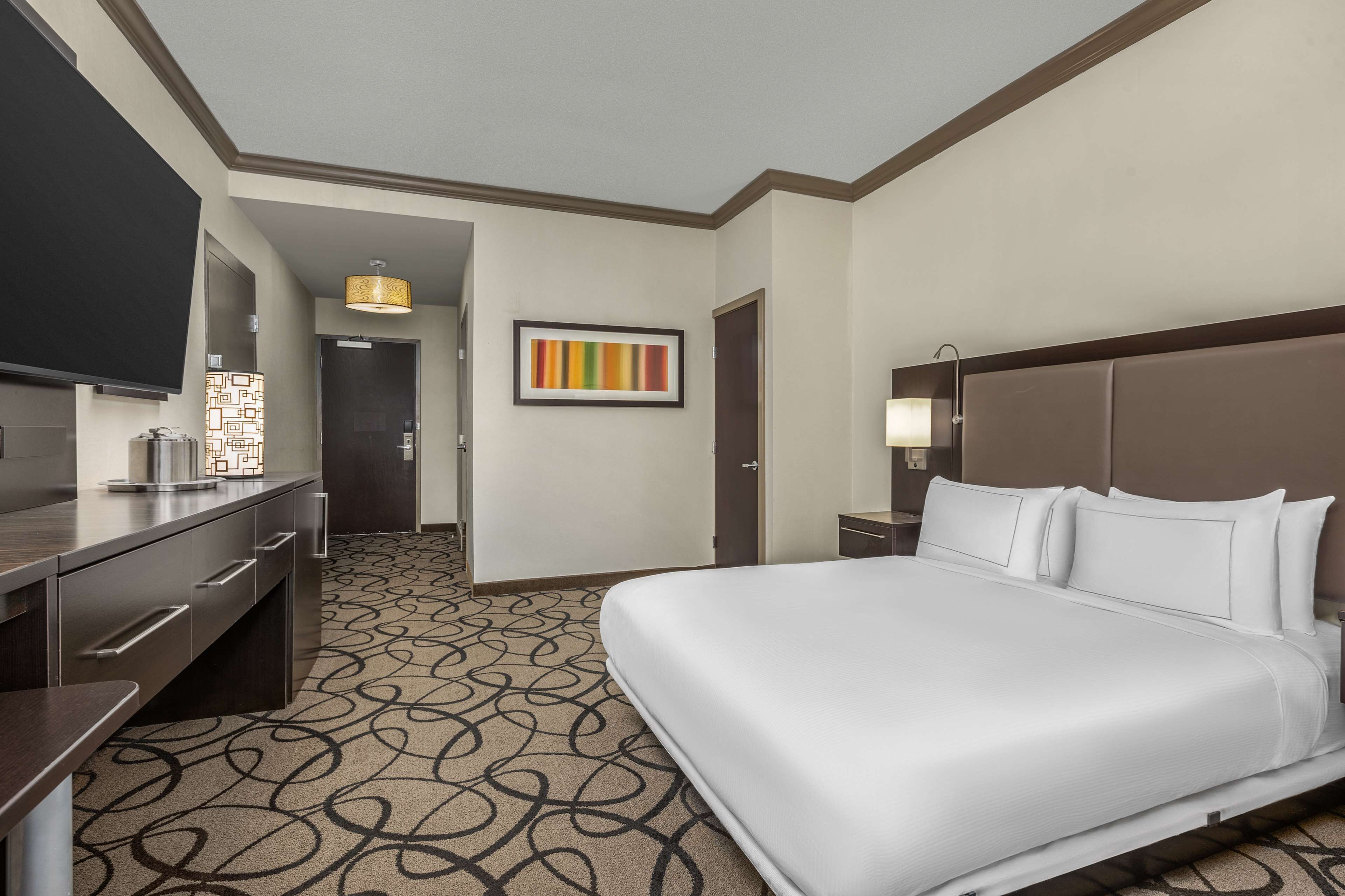 A hotel room with a large bed, a desk with drawers, a flat-screen TV, a patterned carpet, and a framed artwork on the wall.