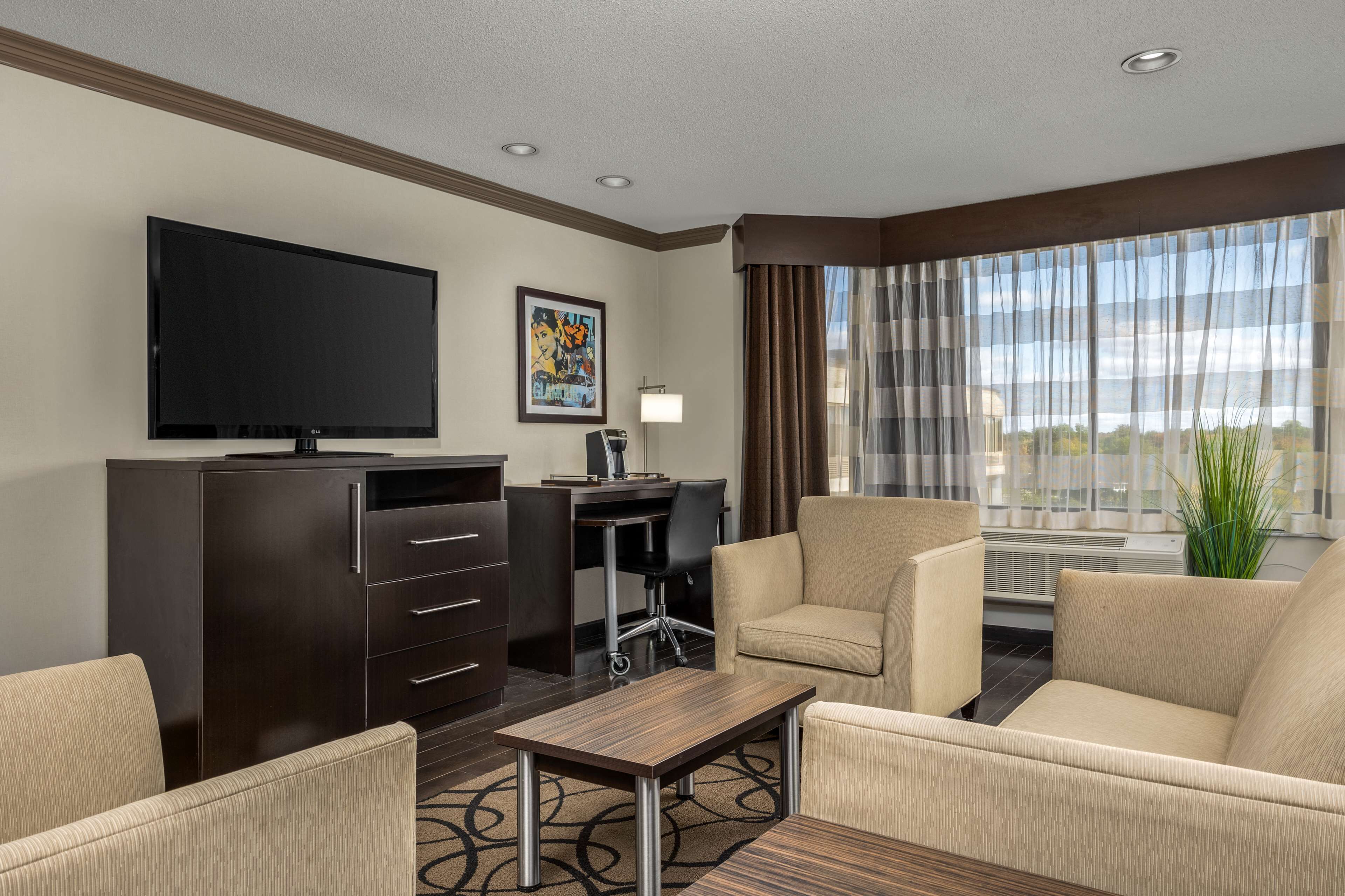 A hotel room with a flat-screen TV, a desk with a chair, a sofa, and a large window with curtains.