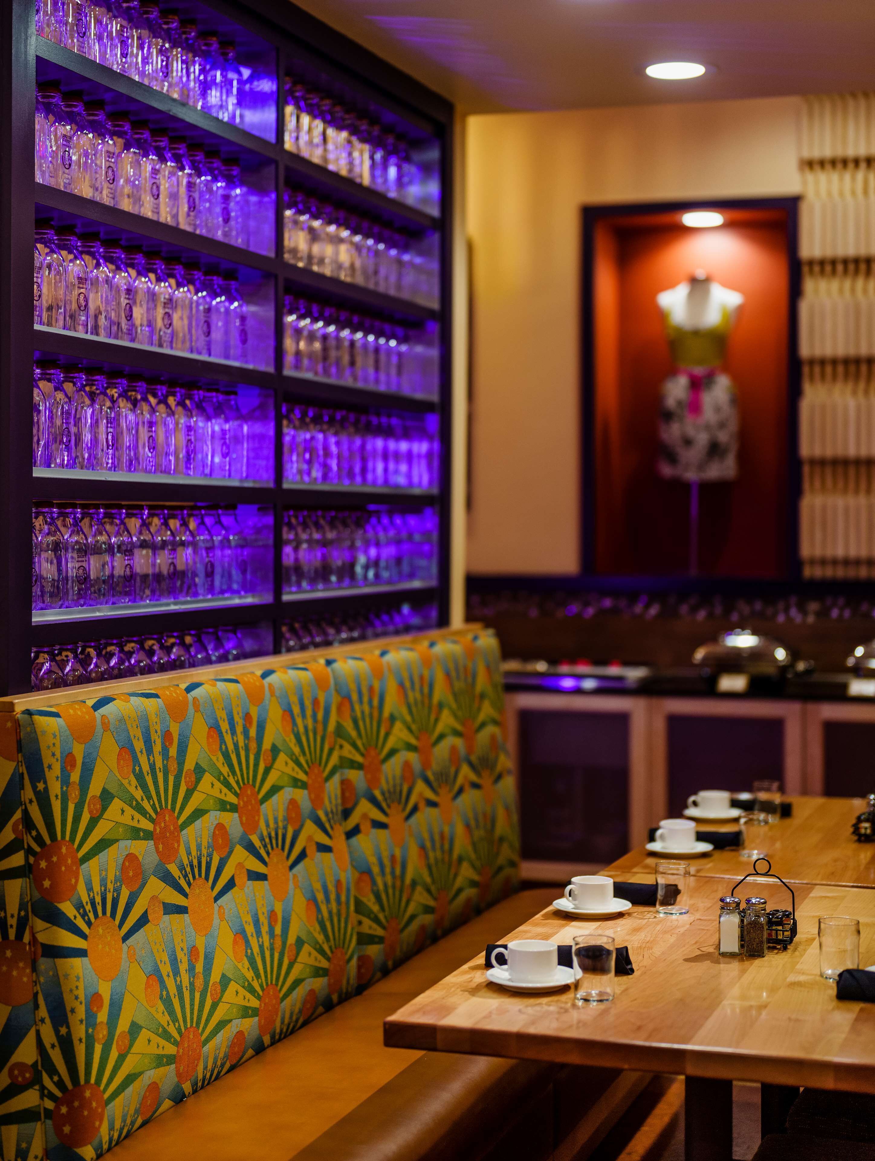 A restaurant with a long wooden table, a patterned bench, and shelves of glasses.