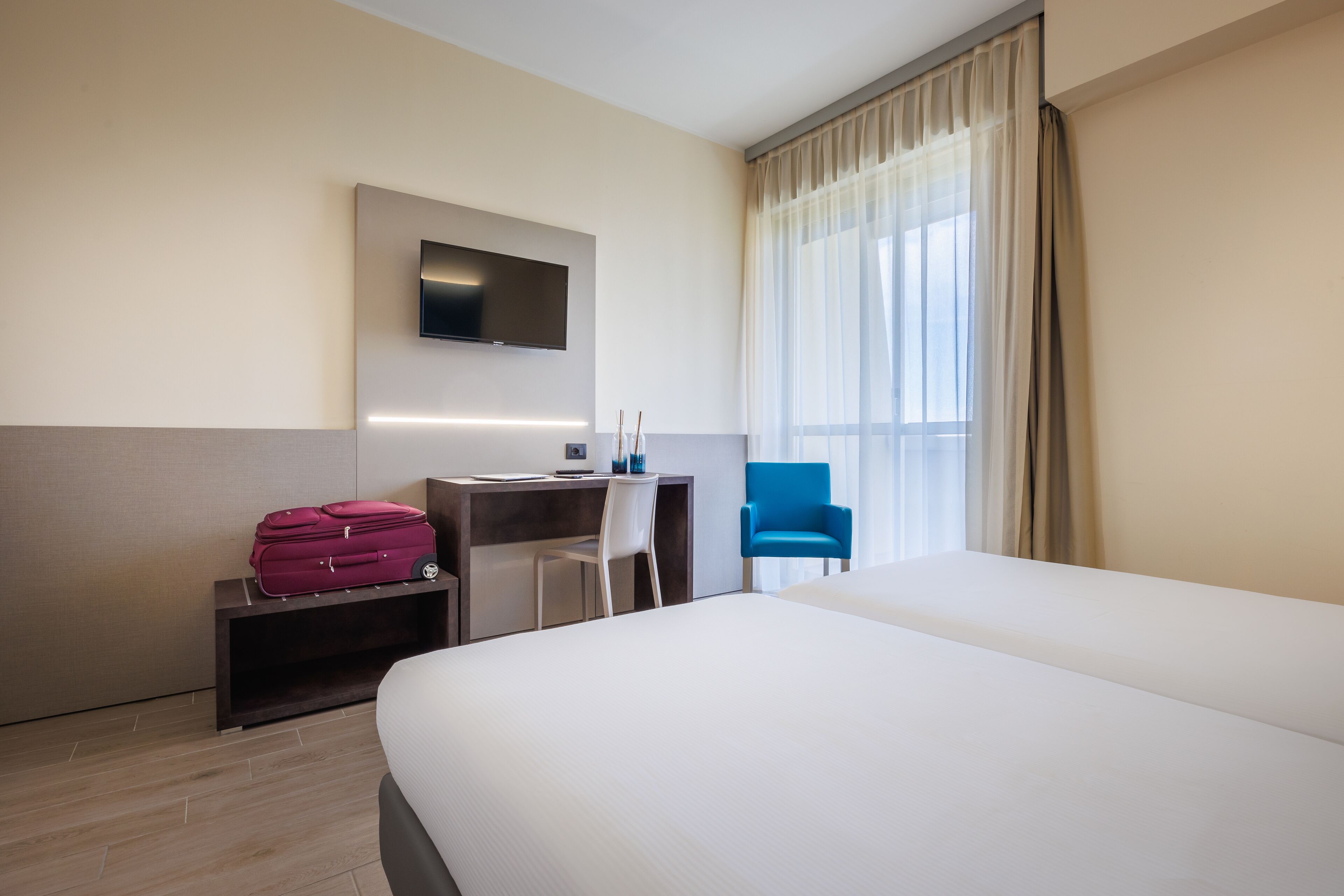 Habitación de hotel con una cama grande, un escritorio con computadora, una silla, televisor montado en la pared y una ventana con cortinas.