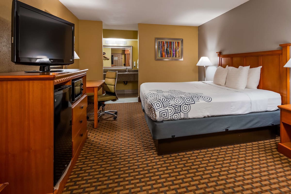 A hotel room with a bed, bedside lamps, a TV, a desk with a chair, and a bathroom visible through an open door.