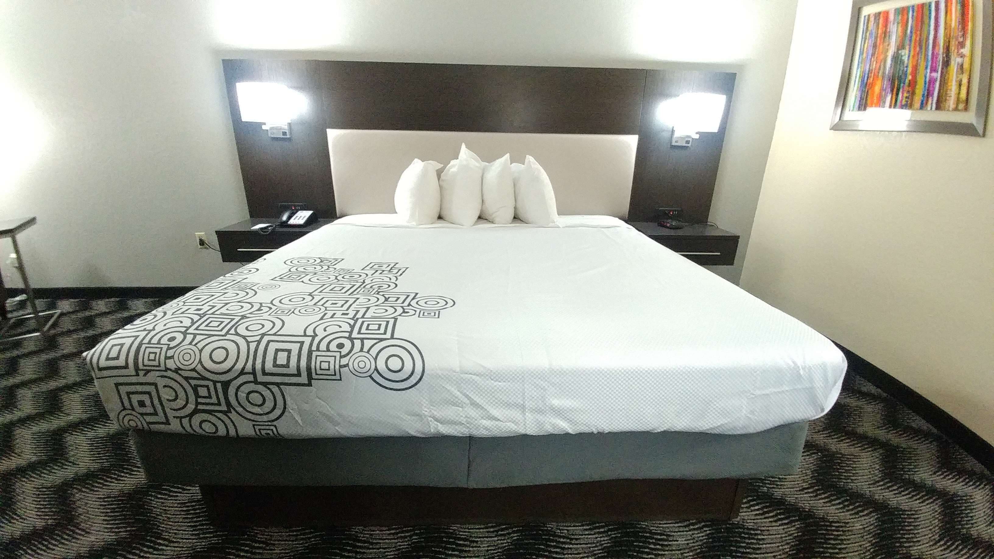 A hotel room with a large bed, a headboard with lights, a desk with a phone, and a wall with a colorful artwork.