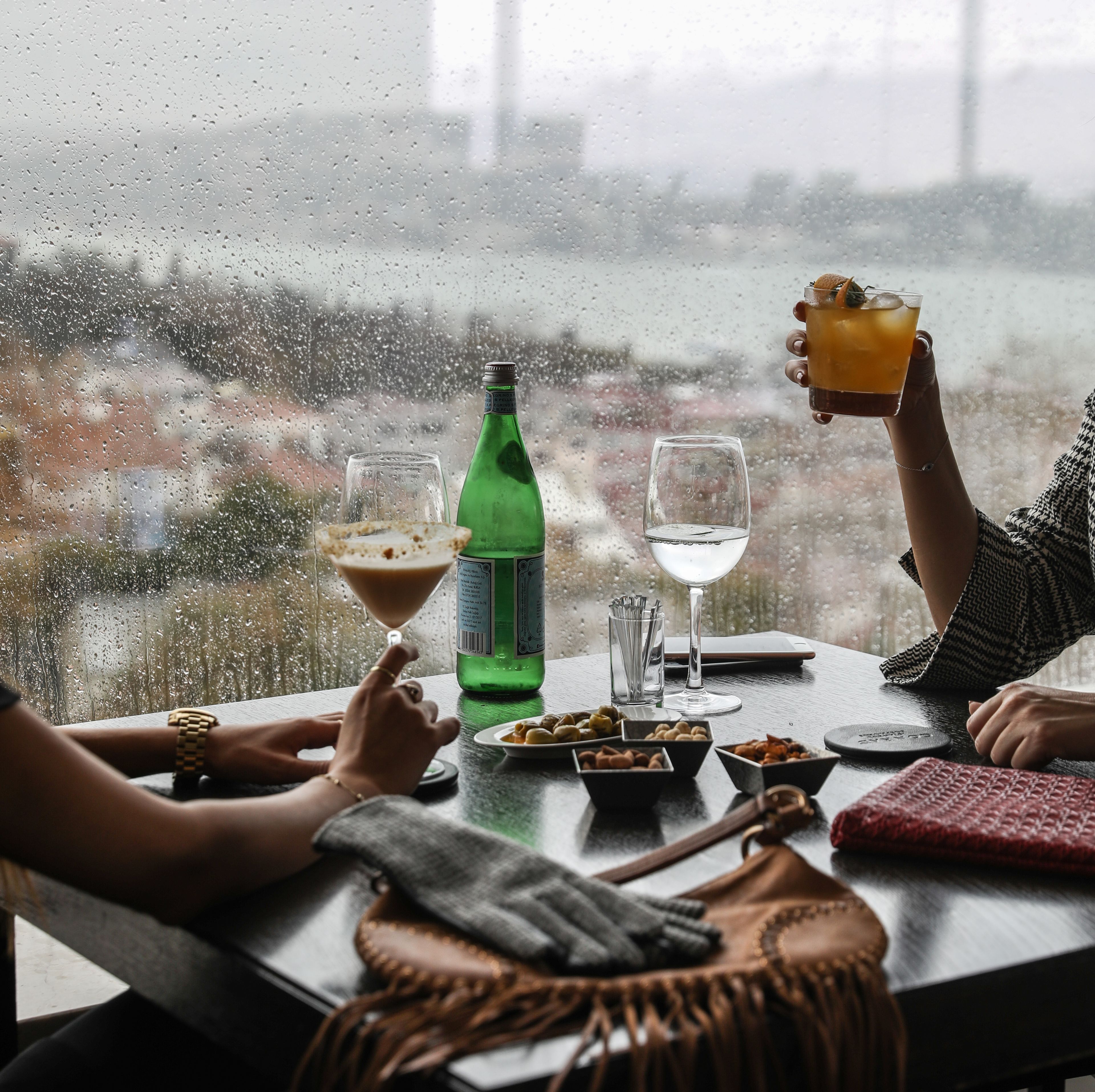Escena de día lluvioso con una mesa dispuesta para dos, que incluye una botella de refresco verde, dos vasos y una copa de martini con una bebida.
