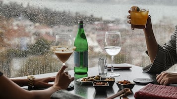 Escena de día lluvioso con una mesa dispuesta para dos, que incluye una botella de refresco verde, dos vasos y una copa de martini con una bebida.