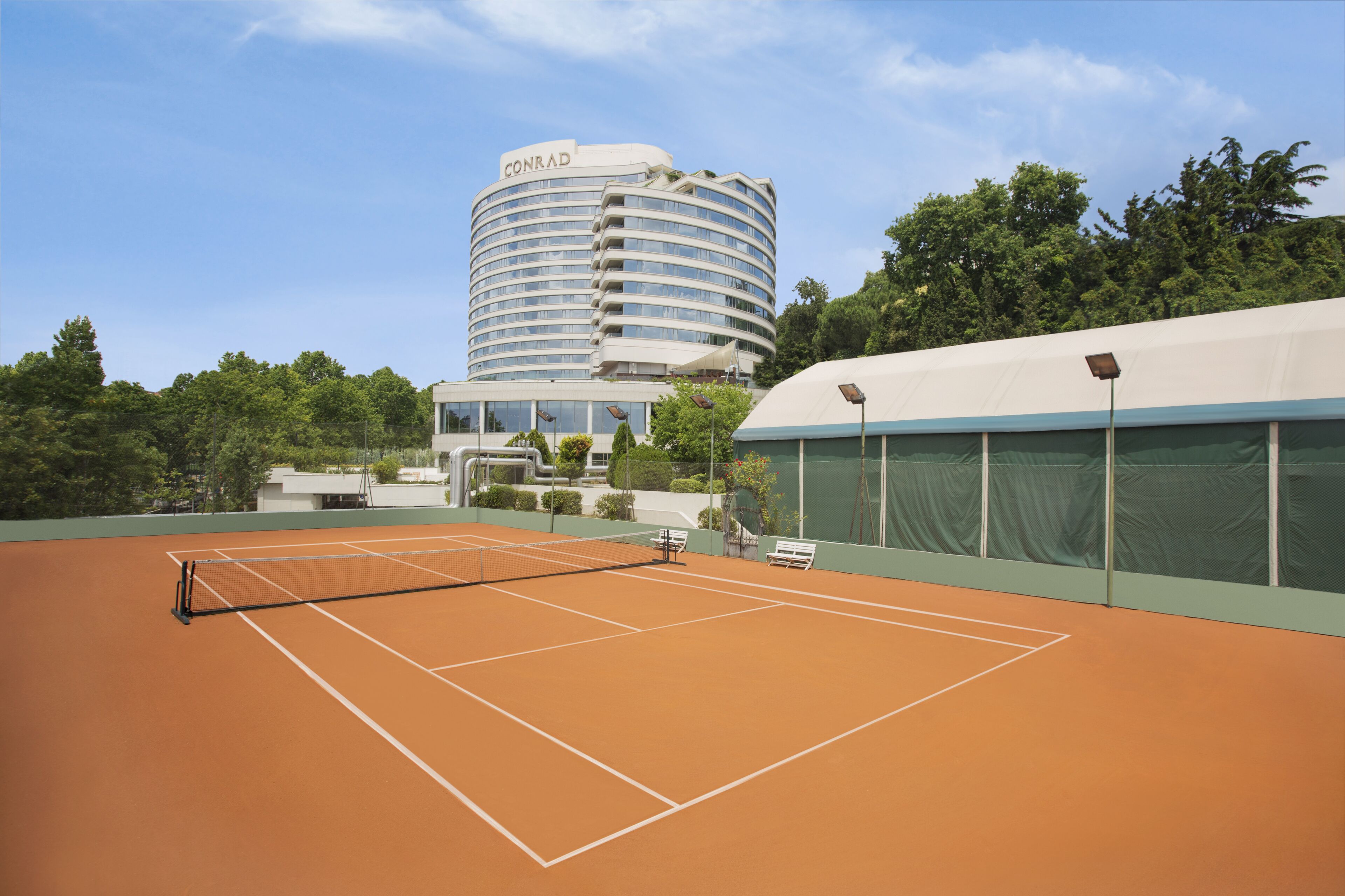 Un campo da tennis con una rete, panchine e un edificio con la scritta 'CONRAD' sullo sfondo.
