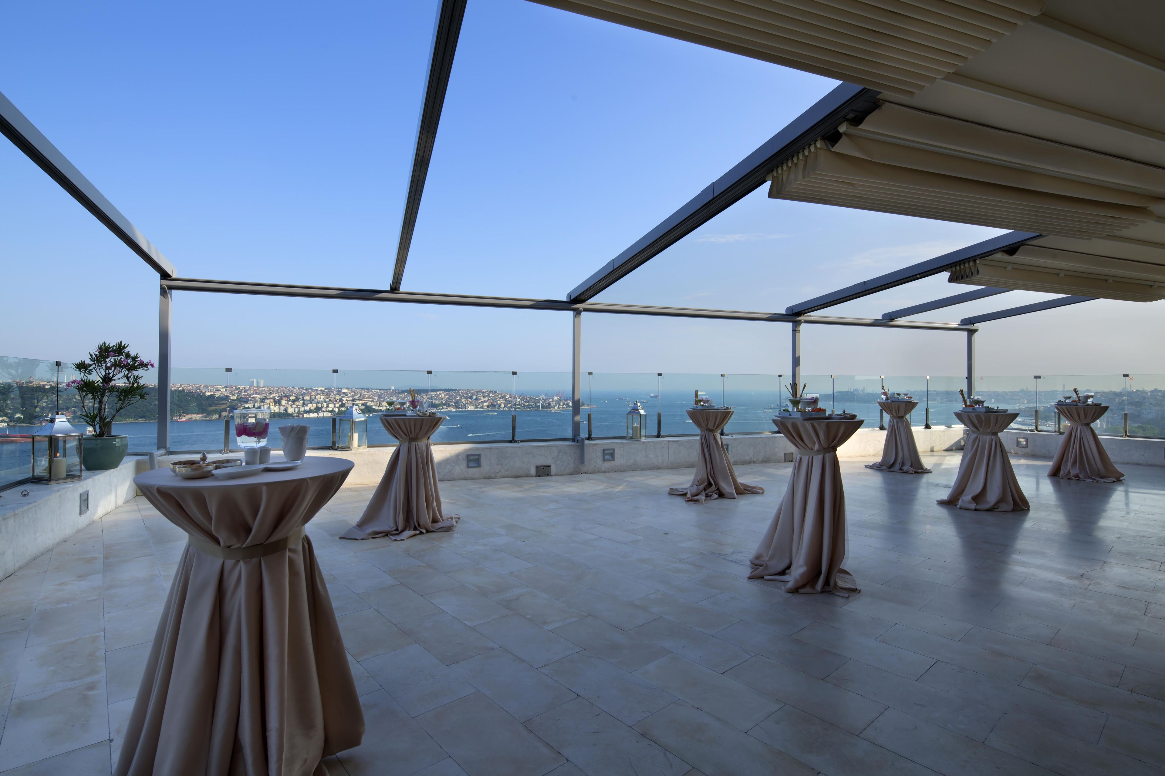 A terrace with tables set for dining, overlooking a cityscape and the sea.