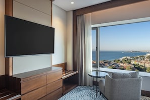 A modern hotel room with a large TV, a small table, a comfortable chair, and a view of the sea and city. - Conrad Istanbul Bosphorus (Istanbul)