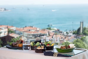 A table with various dishes and glasses, overlooking a coastal cityscape with buildings and the sea. - Conrad Istanbul Bosphorus (Istanbul)