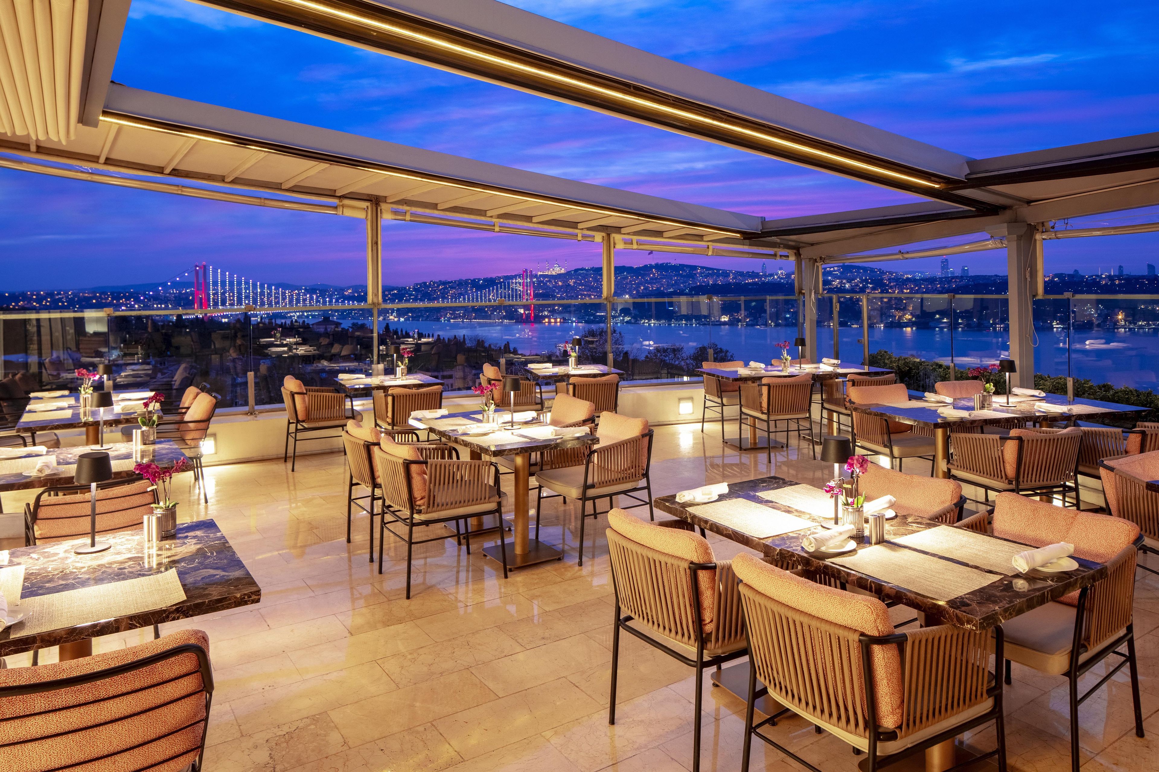 A rooftop restaurant with tables and chairs, a city view, and a glass ceiling.