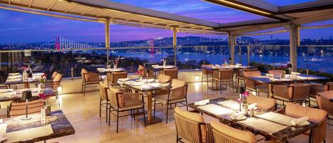 A rooftop restaurant with tables and chairs, a city view, and a glass ceiling.