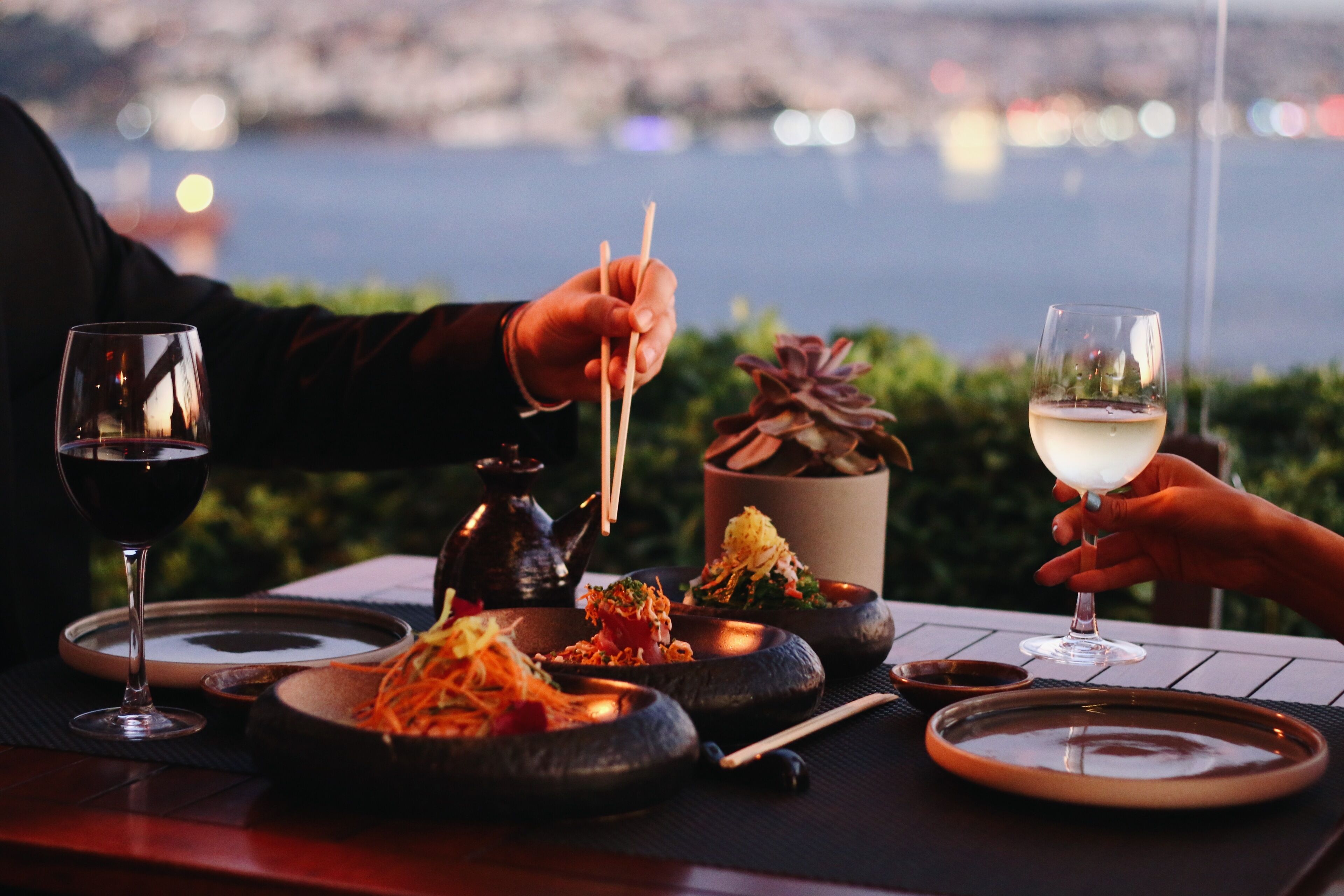 A table set with dishes and drinks, a person holding chopsticks, and a glass of white wine.