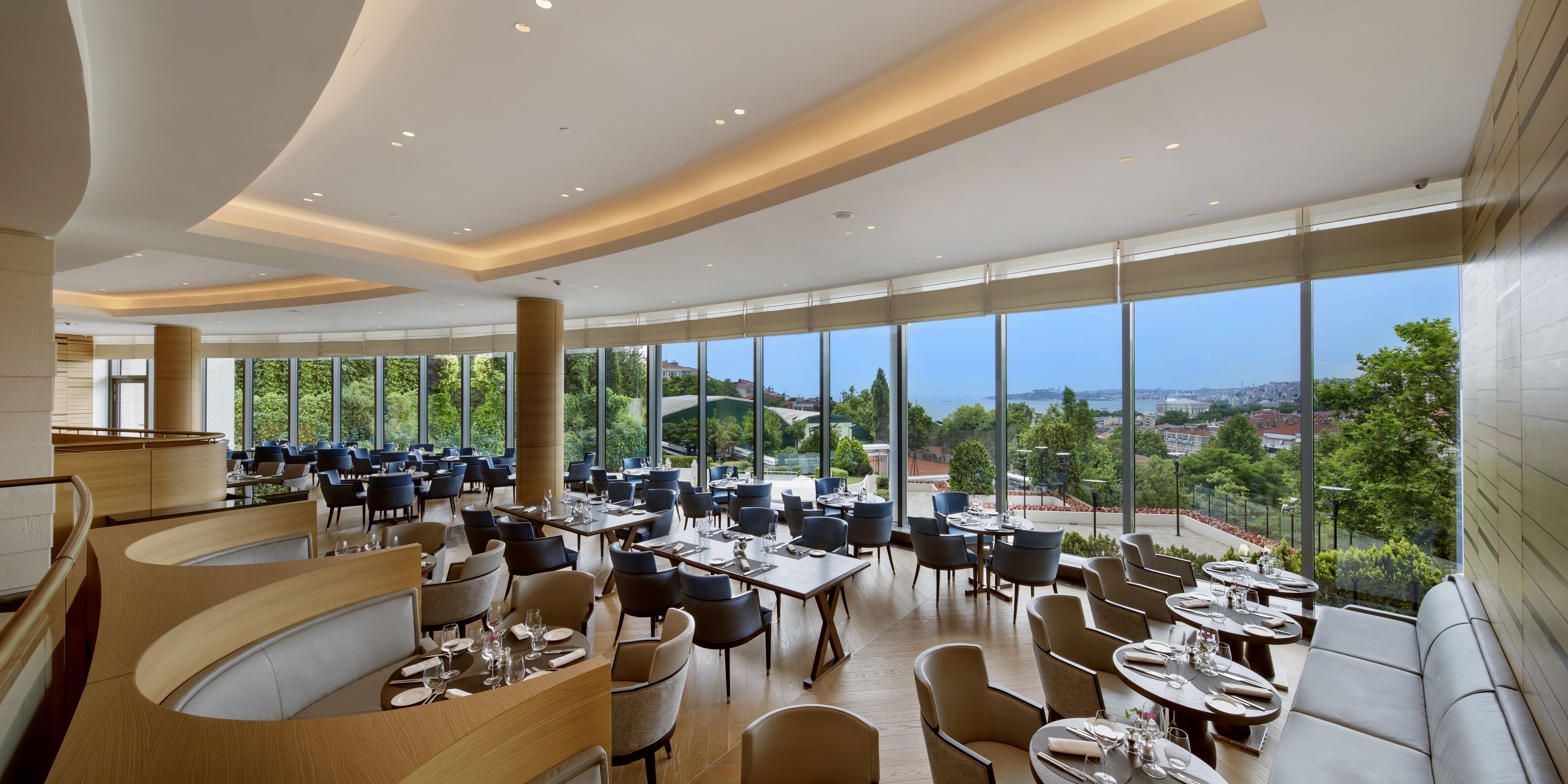 A spacious restaurant with multiple dining areas, large windows offering a city view, and a modern interior design.