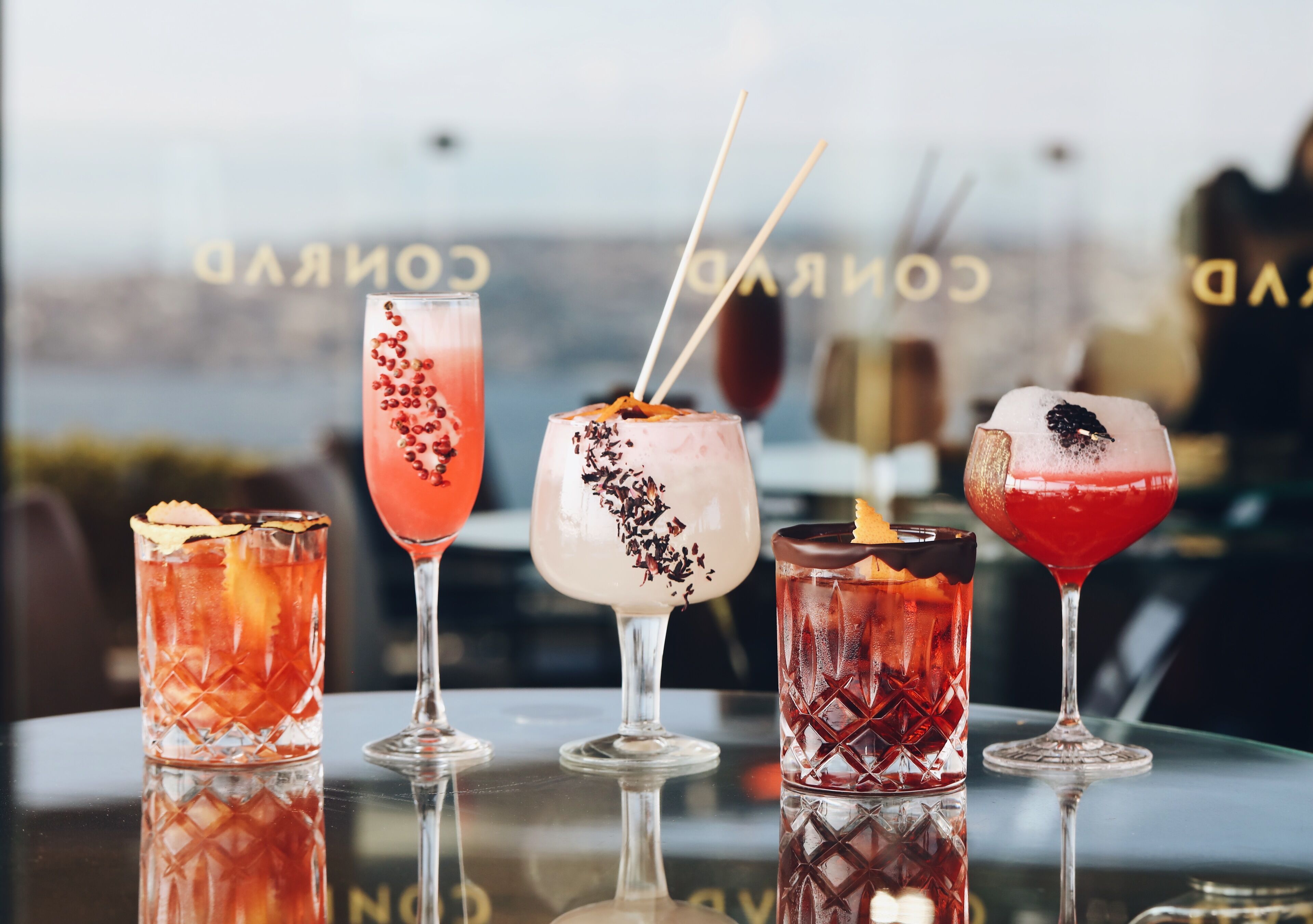 Vier Cocktails auf einer spiegelnden Oberfläche, im Hintergrund verschwommen der Schriftzug 'CORAL'.