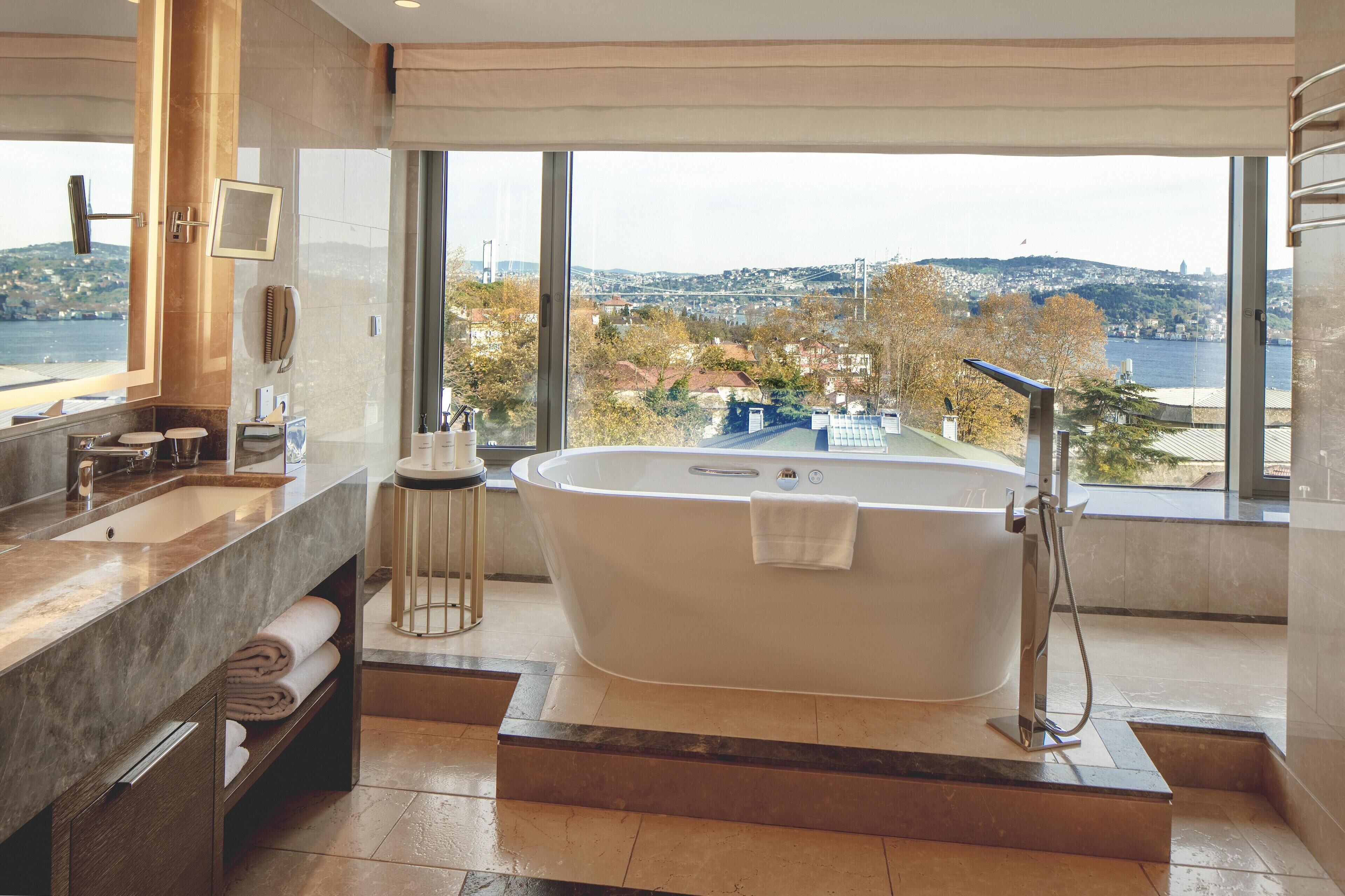 A modern bathroom with a large bathtub, a vanity with a sink, and a large window offering a view of a cityscape and a bridge.