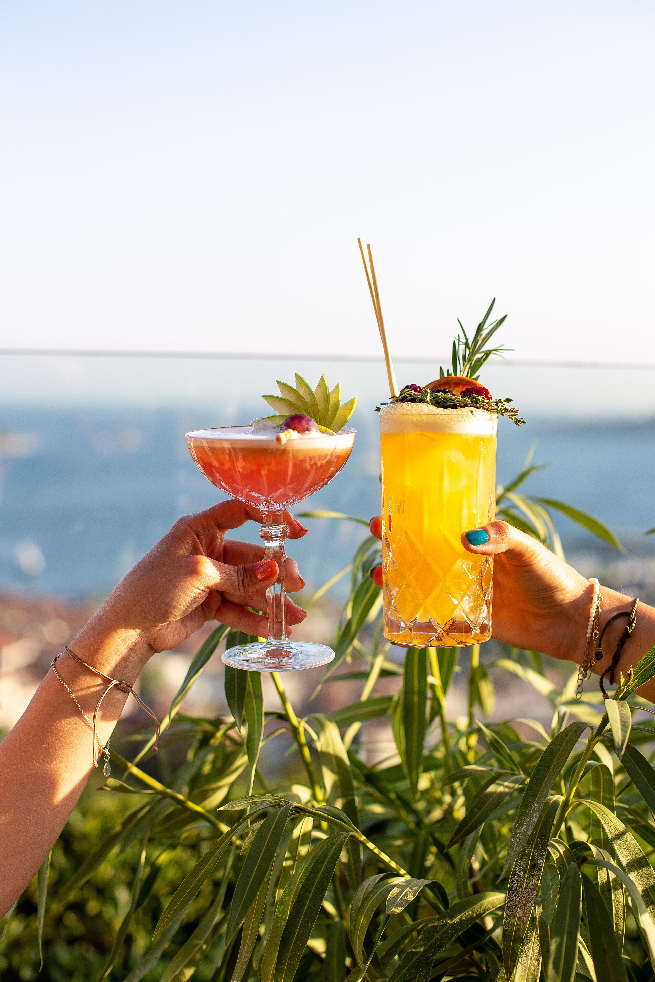Duas mãos segurando copos de coquetel com uma bebida tropical, com um fundo de vegetação e uma vista distante.
