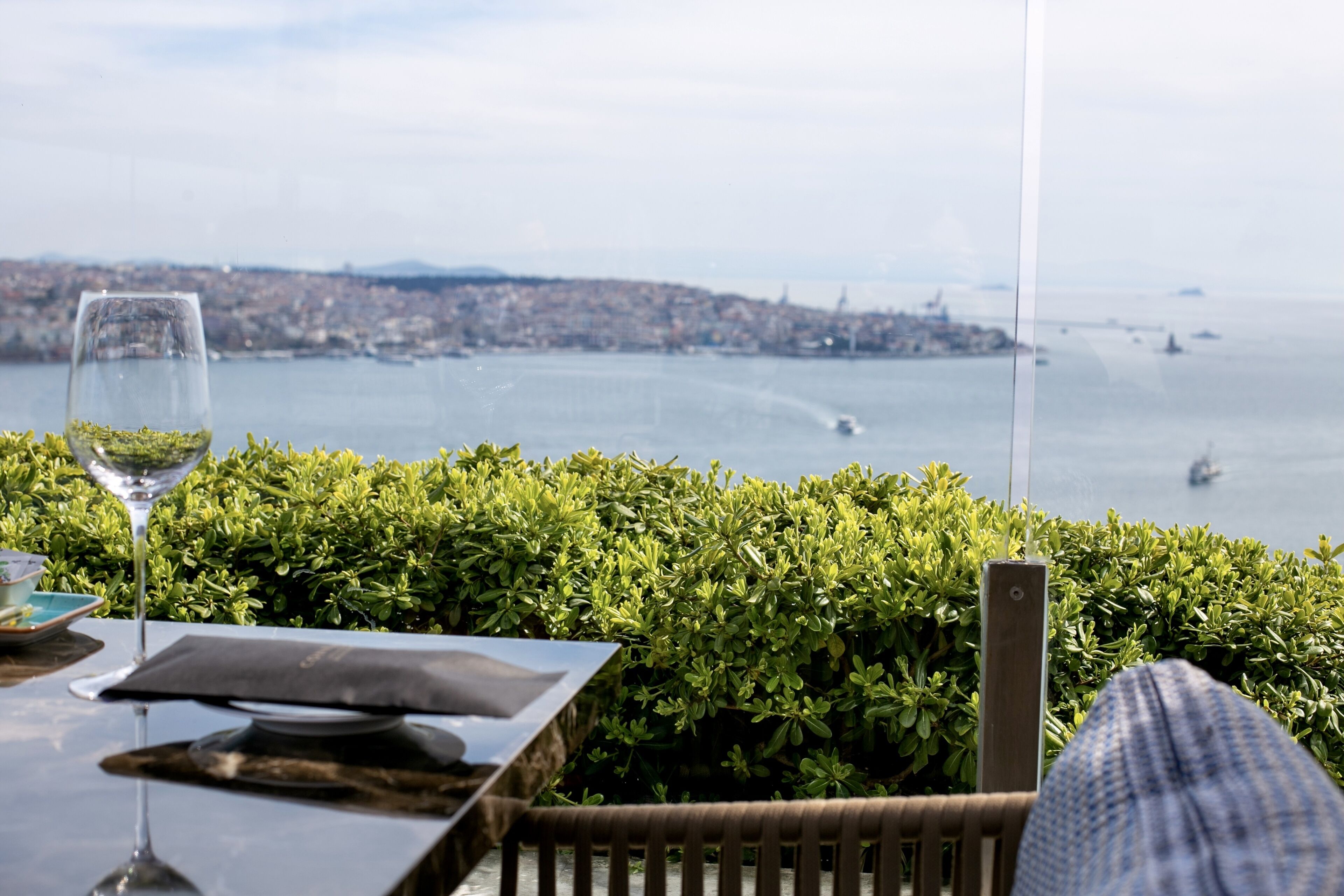 A table with a glass of water, a menu, and a napkin, overlooking a body of water with boats and a distant cityscape.