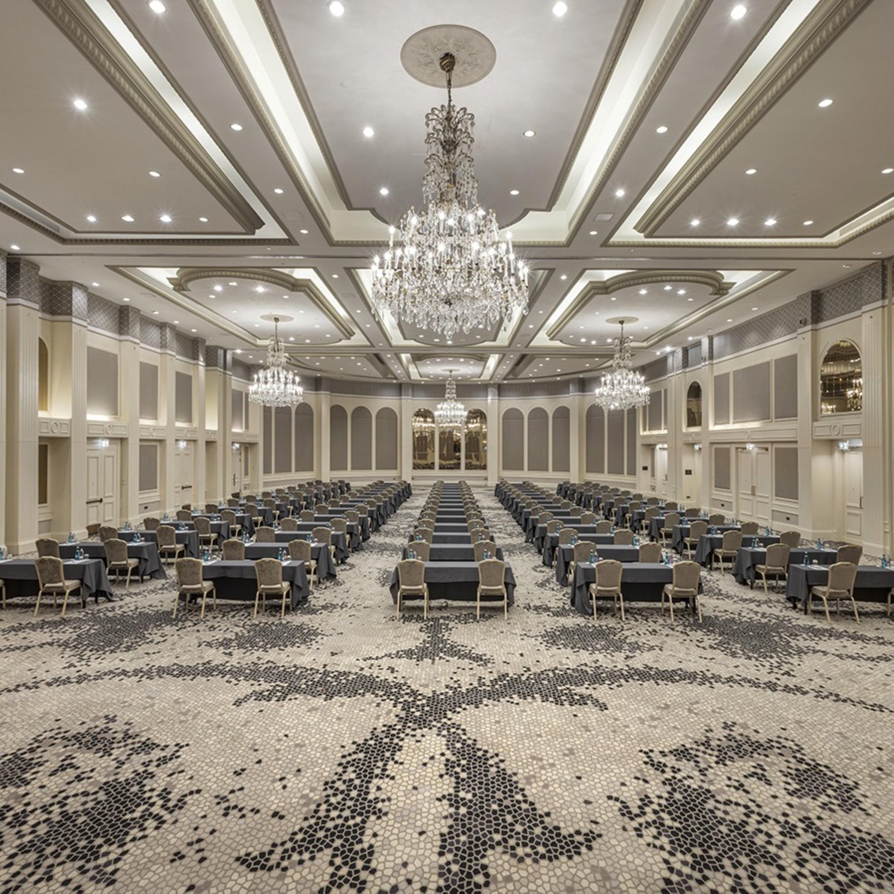 A spacious conference room with rows of tables and chairs, a patterned carpet, and a large chandelier.