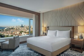 A hotel room with a large bed, a chair, a nightstand, and a view of a cityscape. - Conrad Istanbul Bosphorus (Istanbul)