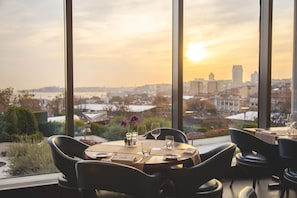 A dining area with a table set for two, overlooking a cityscape with a sunset view.
