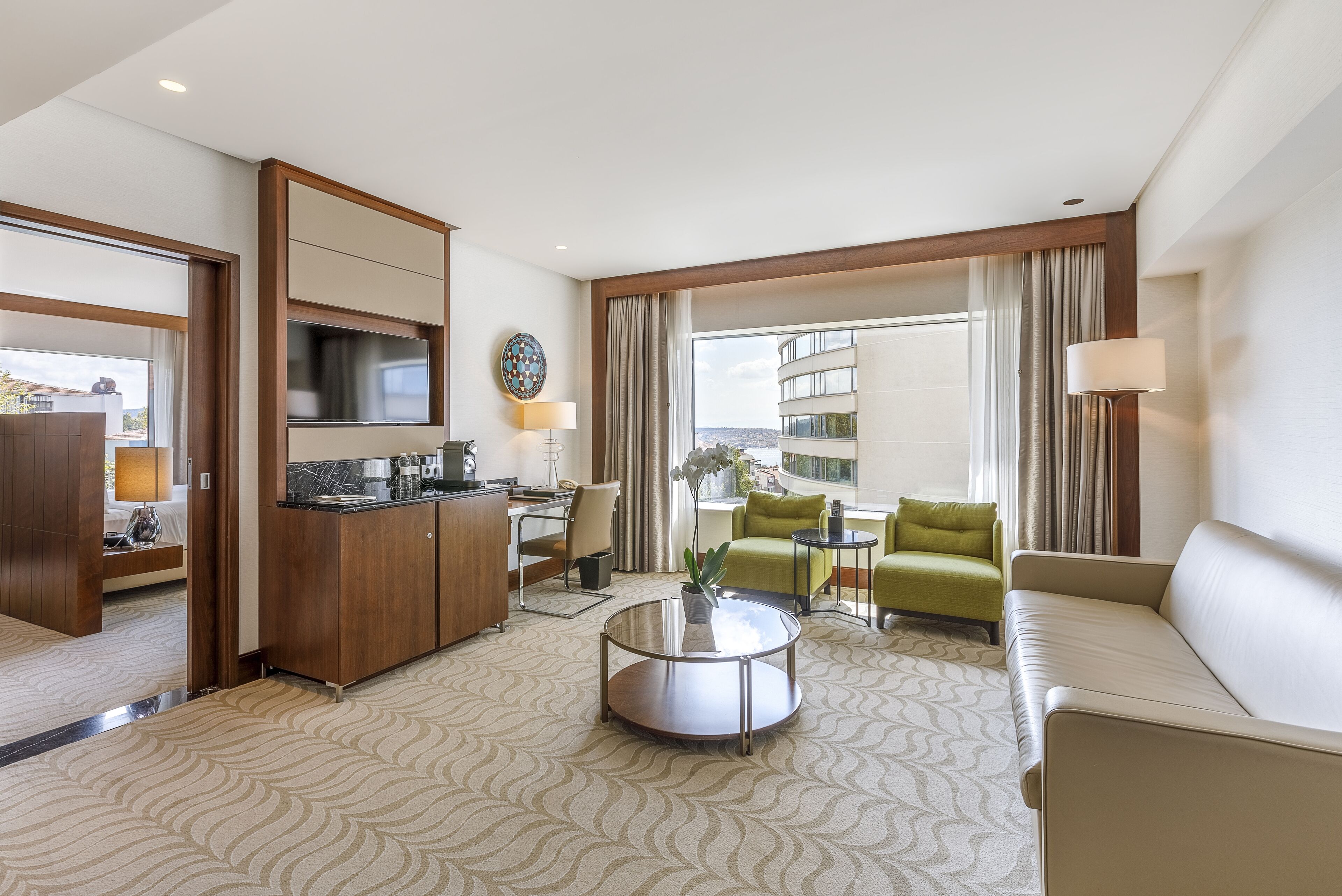 A hotel room with a kitchenette, a round glass coffee table, a sofa, a green armchair, and a view of a cityscape.