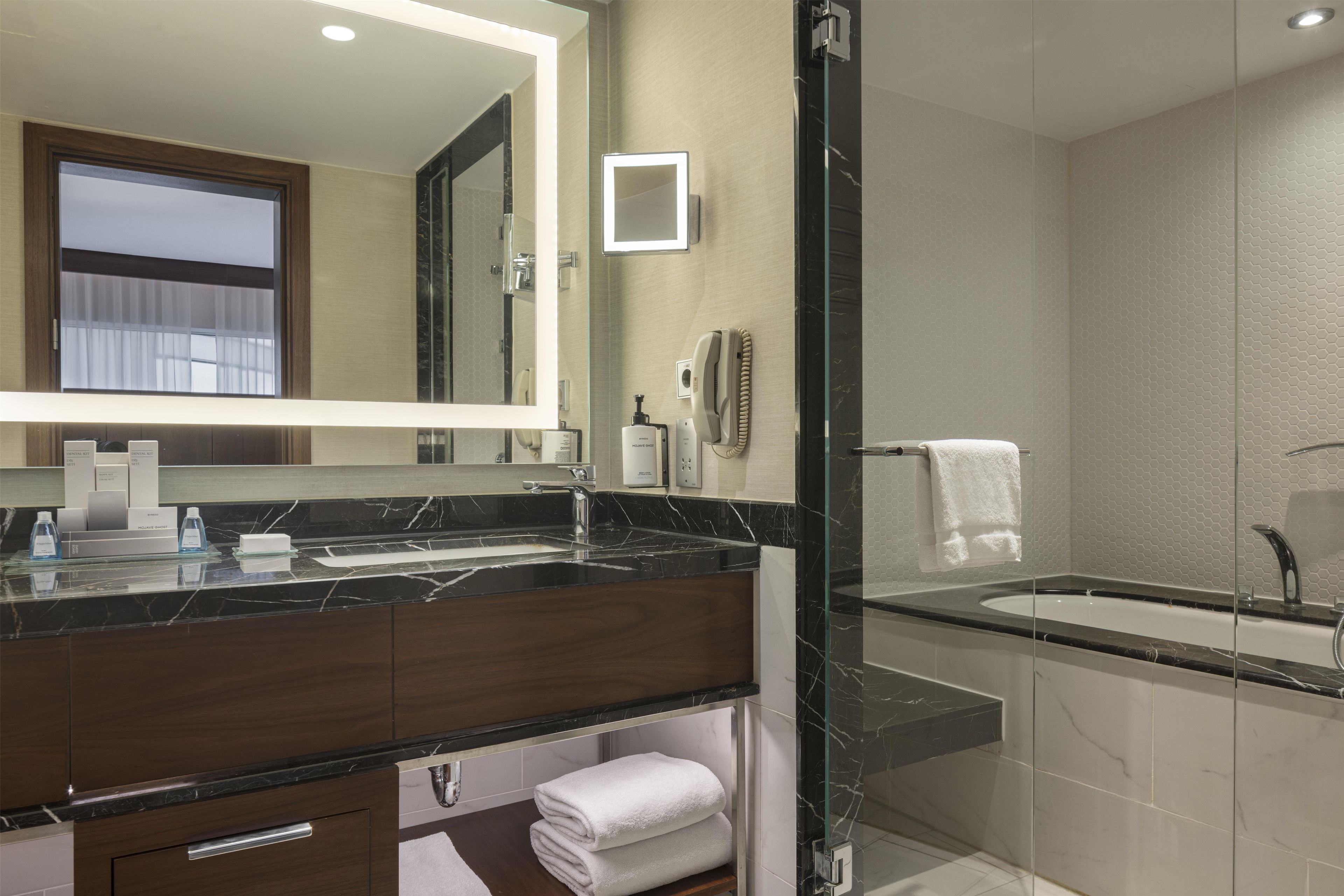 A modern bathroom with a marble countertop, a large mirror, a glass-enclosed shower, and a visible toilet paper dispenser.