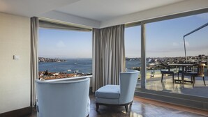 A room with a large window offering a city and sea view, two armchairs, and a table with chairs on an outdoor balcony.