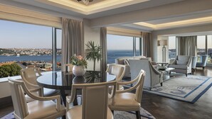 Ein modernes Hotelzimmer mit großem Fenster und Blick auf die Stadt, einem runden Esstisch mit Stühlen, einer Couch und einem gemusterten Teppich.