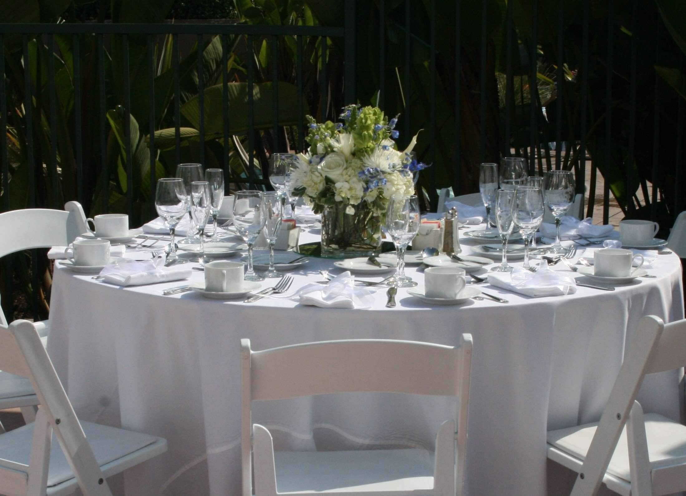 A round table set for a formal outdoor event with white tablecloths, glassware, and a floral centerpiece.