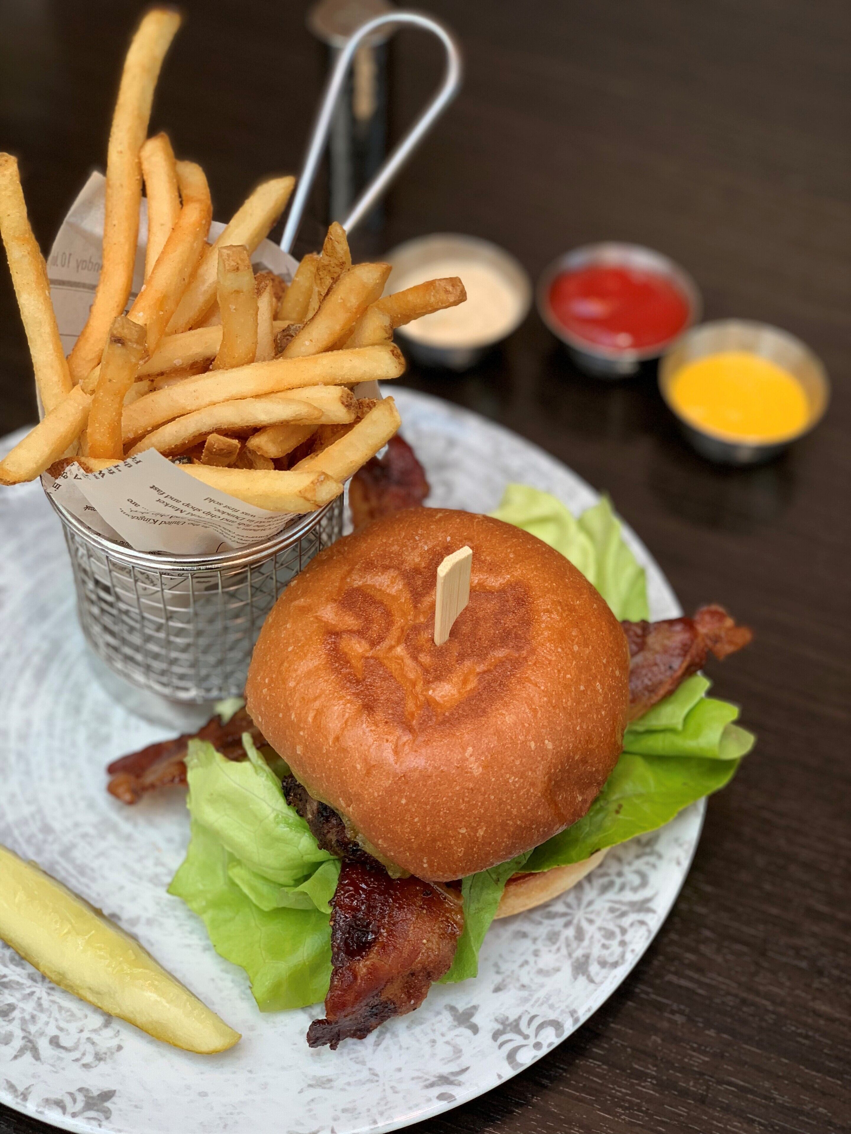 Hamburguesa con tocino, lechuga y pan, acompañada de papas fritas y salsas.