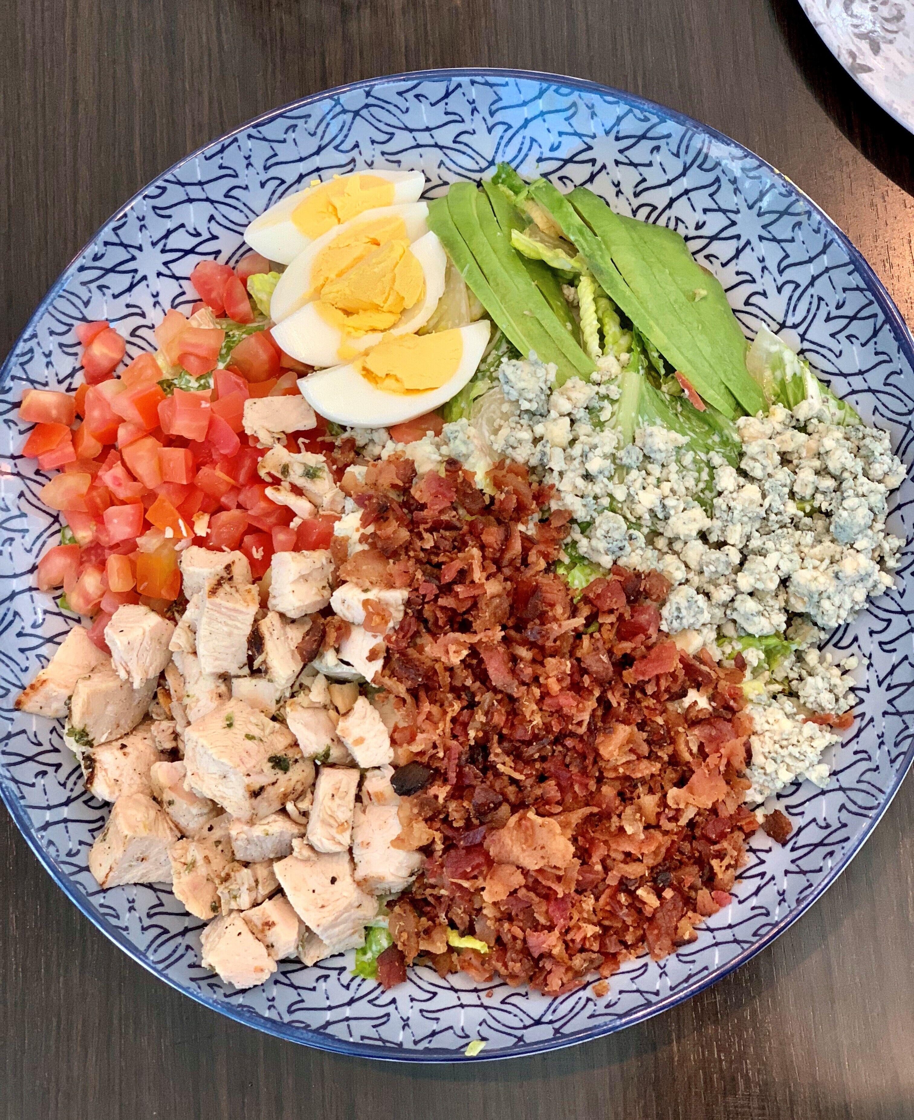 Une assiette contenant divers aliments, notamment des tomates coupées en dés, du poulet en tranches, des lardons, des œufs durs et une salade composée de laitue et de fromage bleu.