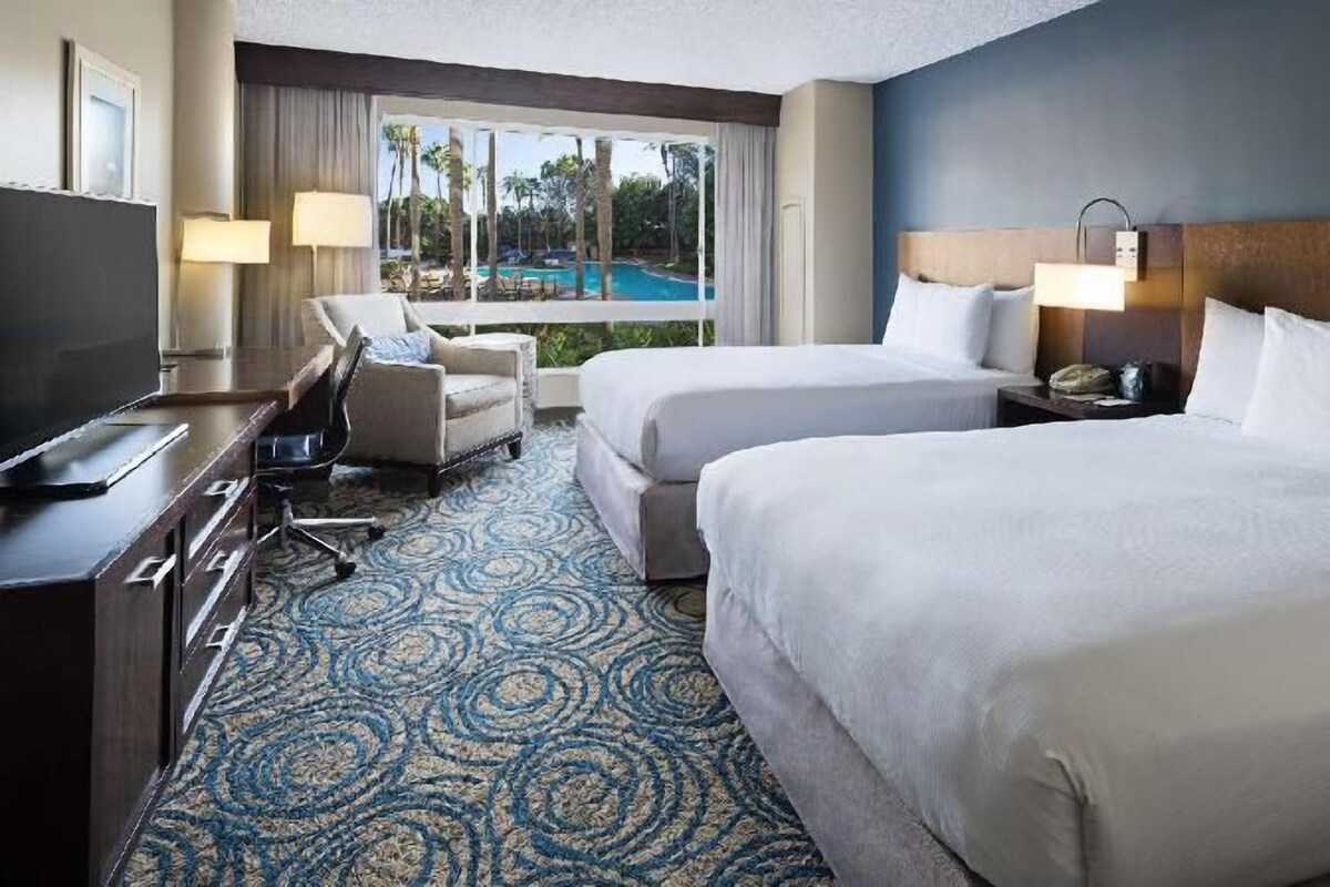 A hotel room with two beds, a desk, a chair, a TV, and a view of a pool.
