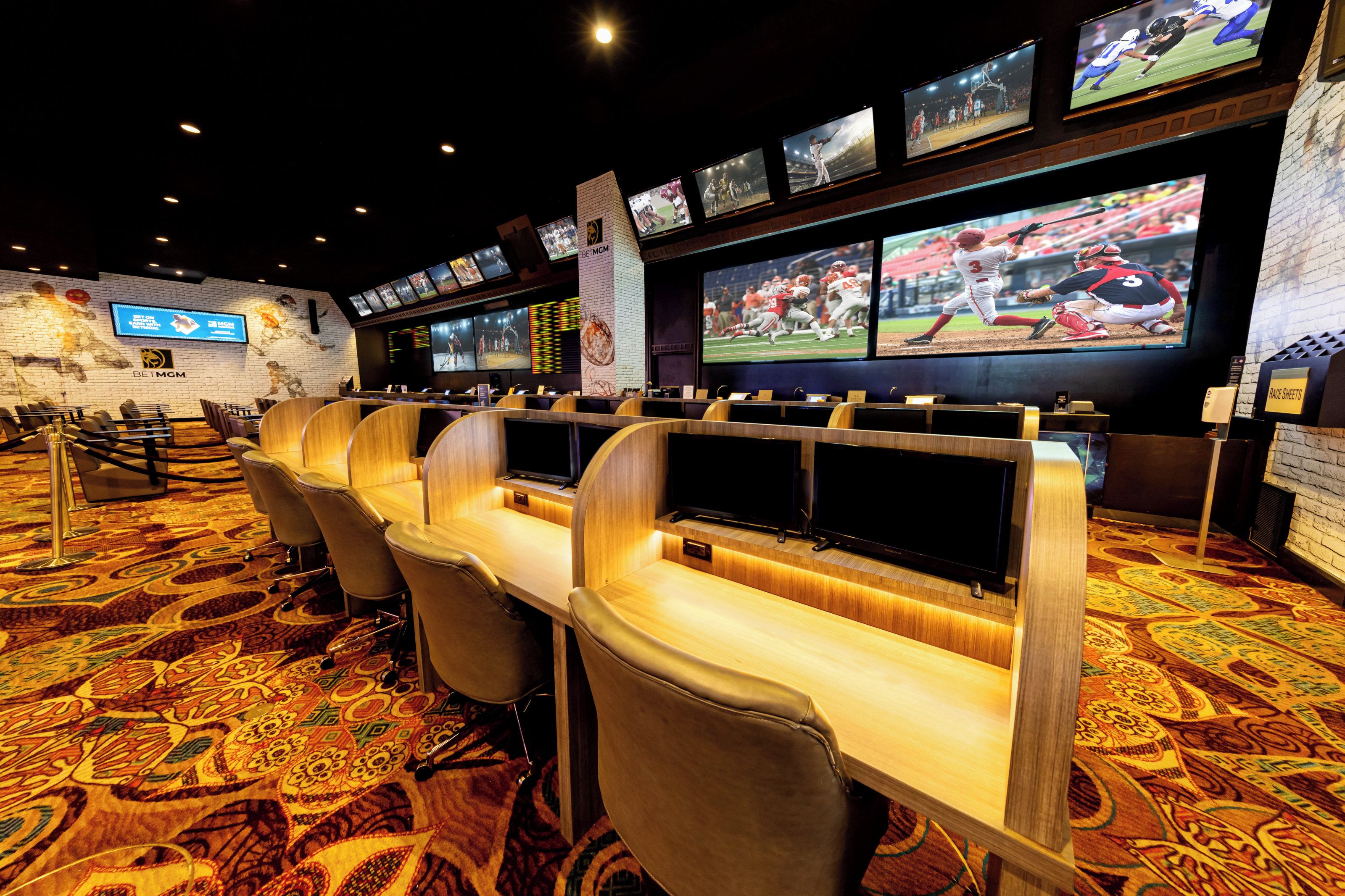 A sports betting area with multiple screens, a brick wall with a large screen, and a carpeted floor.