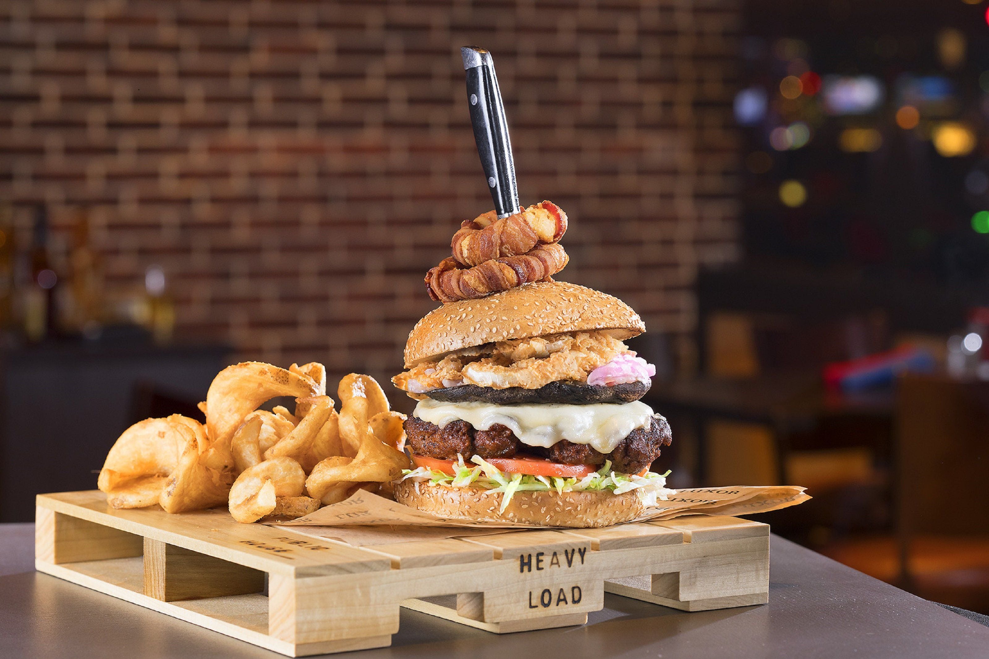 A burger with bacon, cheese, and a pick, served with fries on a wooden board labeled 'HEAVY LOAD'.