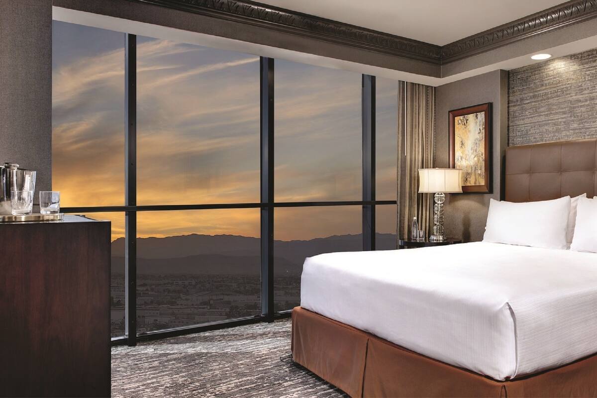Une chambre d’hôtel avec un grand lit, un bureau avec une lampe, une table de chevet et une vue sur le coucher du soleil à travers de grandes fenêtres du sol au plafond.