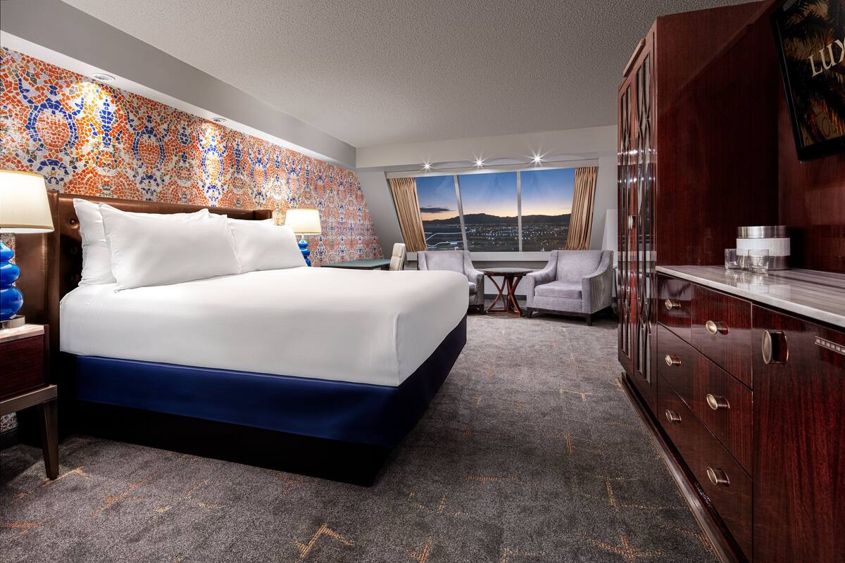 Une chambre d’hôtel avec un grand lit, un bureau avec une lampe, une chaise, une armoire et une vue sur la ville au coucher du soleil.