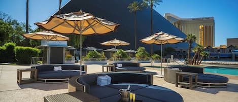 Un área junto a la piscina con sillones de descanso y sombrillas, un edificio con forma de pirámide al fondo y una piscina.