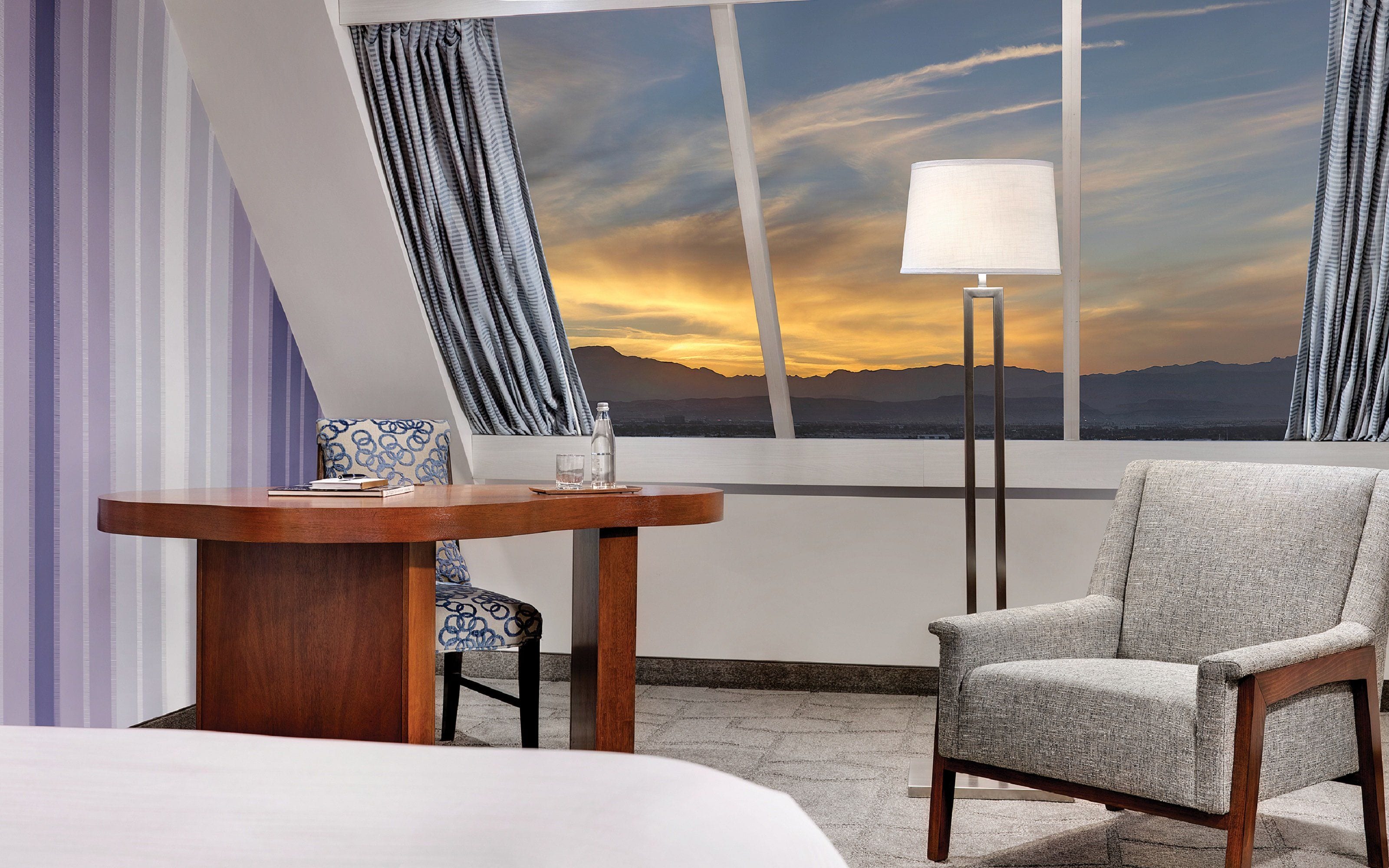 Habitación de hotel con cama, mesa, silla, lámpara y vista a las montañas al atardecer.